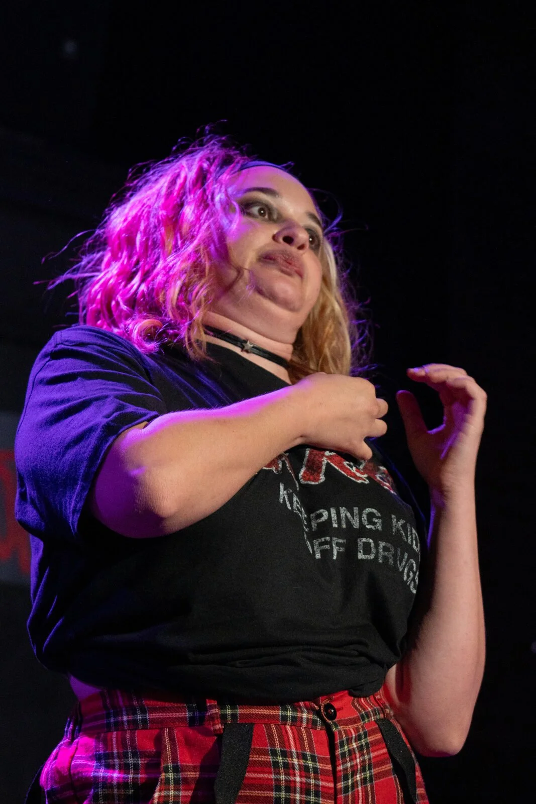 A woman with curly blonde hair, illuminated by pink and purple stage lights, is performing on stage. She wears a black T-shirt and red plaid pants, with her hand raised near her chest.