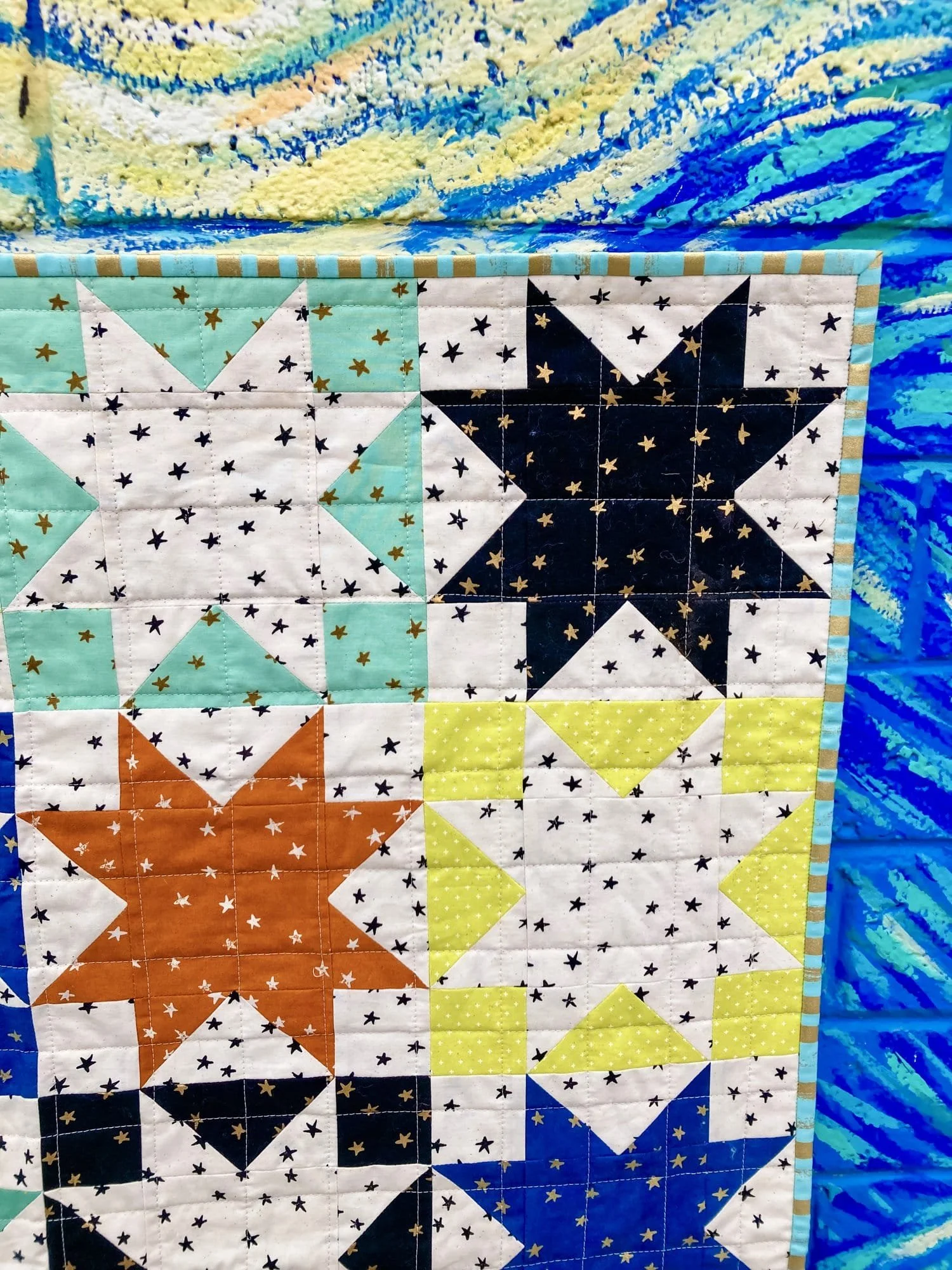 Close-up of a Sawtooth Star quilt with blocks in teal, orange, navy, and yellow, each paired with white fabric printed with small gold stars. The quilt is photographed against a vivid blue and yellow mural background.