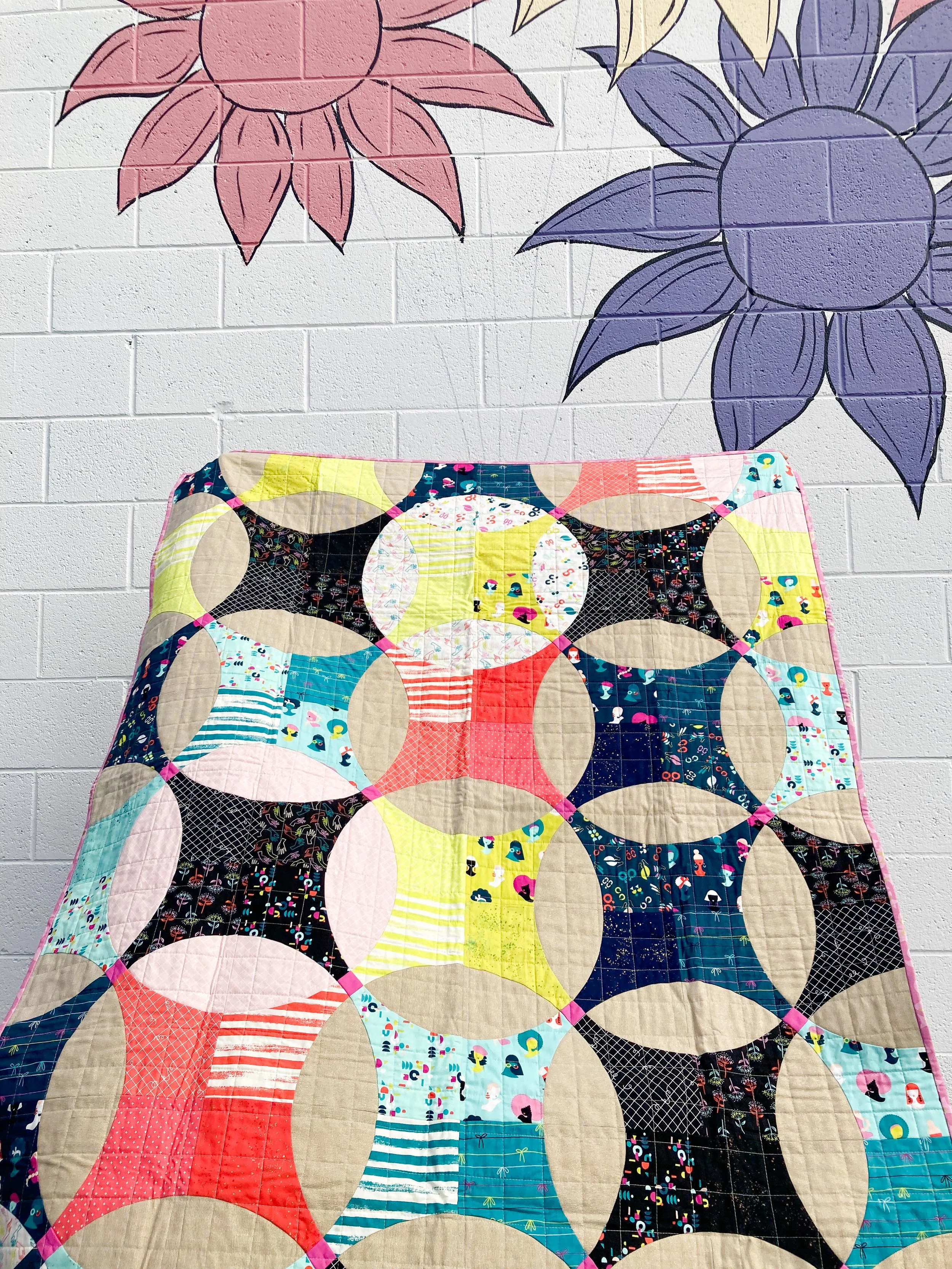 Flowering Snowball quilt made with Ruby Star Society Darlings fabric, featuring bold solids and playful prints in a colorful, modern layout, photographed against a mural with large flowers.