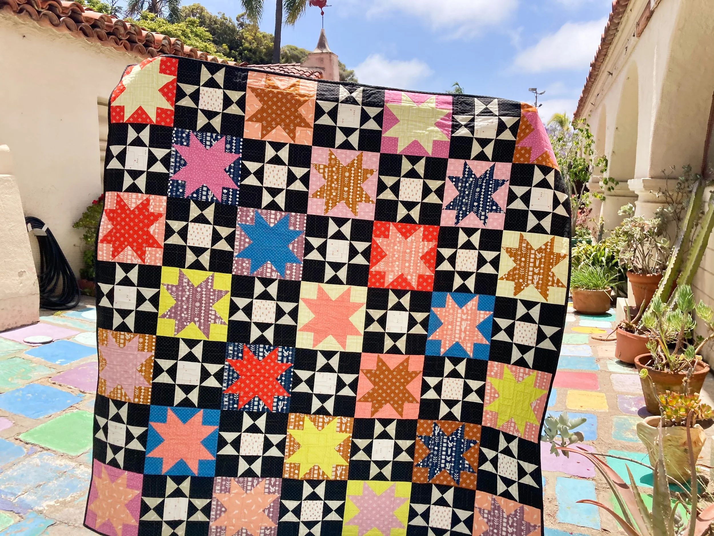 The Mom & Pop quilt featuring Ohio Stars and Sawtooth Stars in a colorful mix of fabrics, displayed outdoors against a tiled patio and mission-style building.