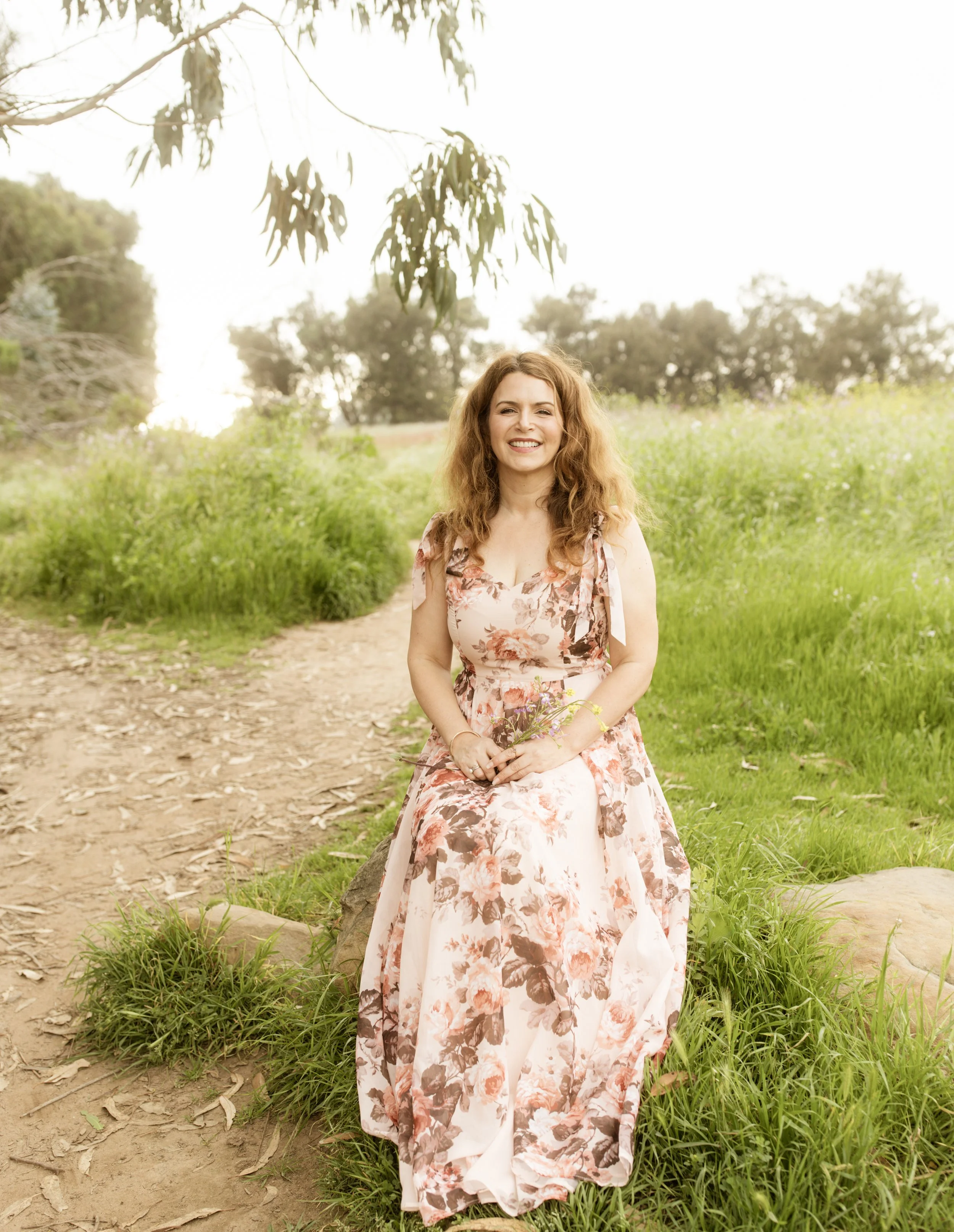 A photo of Leslie Miranda, sitting outside where trees and tufts of grass are visible. She has dark blonde hair, and is wearing a pink floral dress.