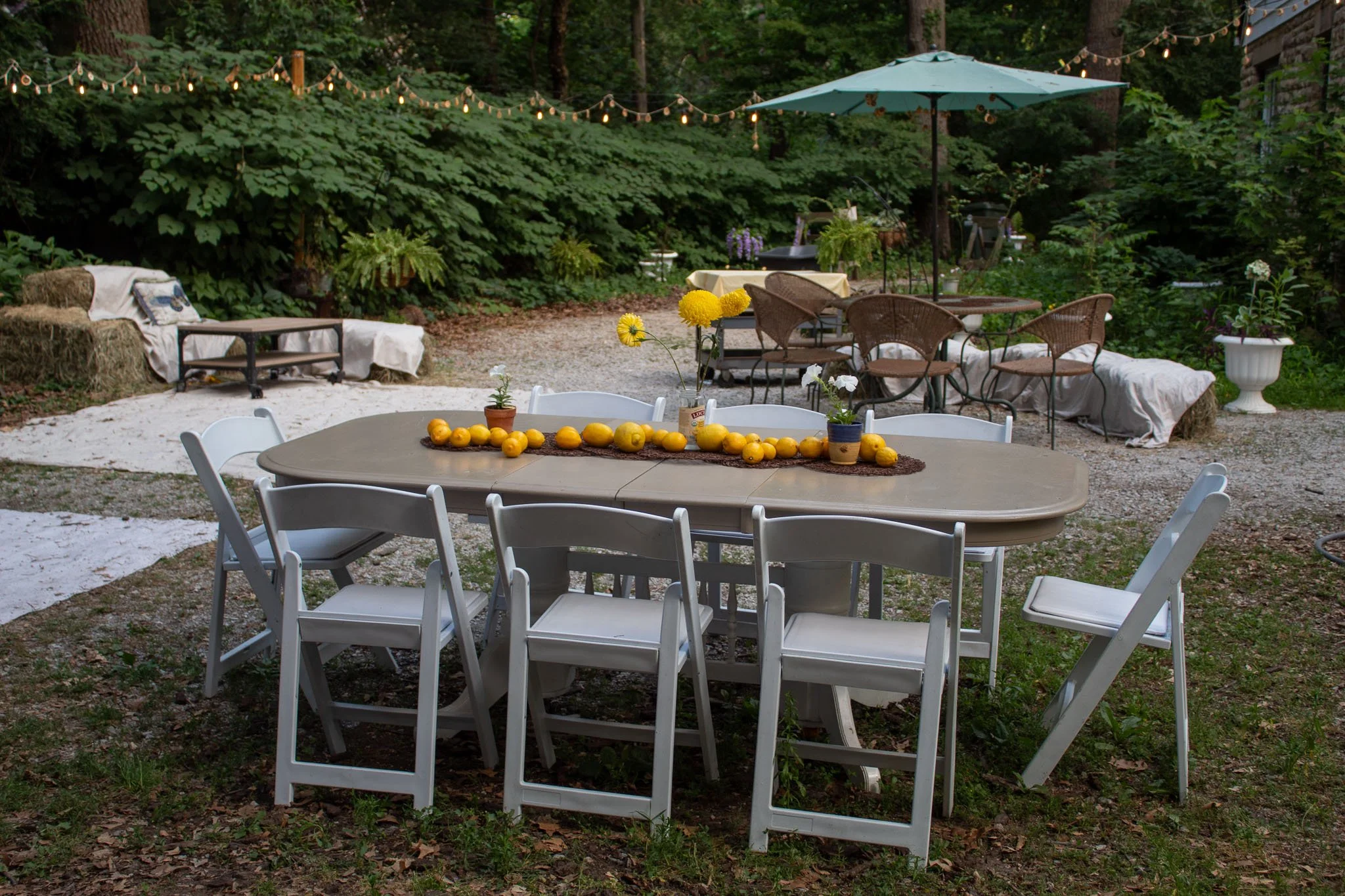 Outdoor garden party seating with white chairs, lemon-decorated tables, hay-bale benches, string lights, and a patio umbrella.