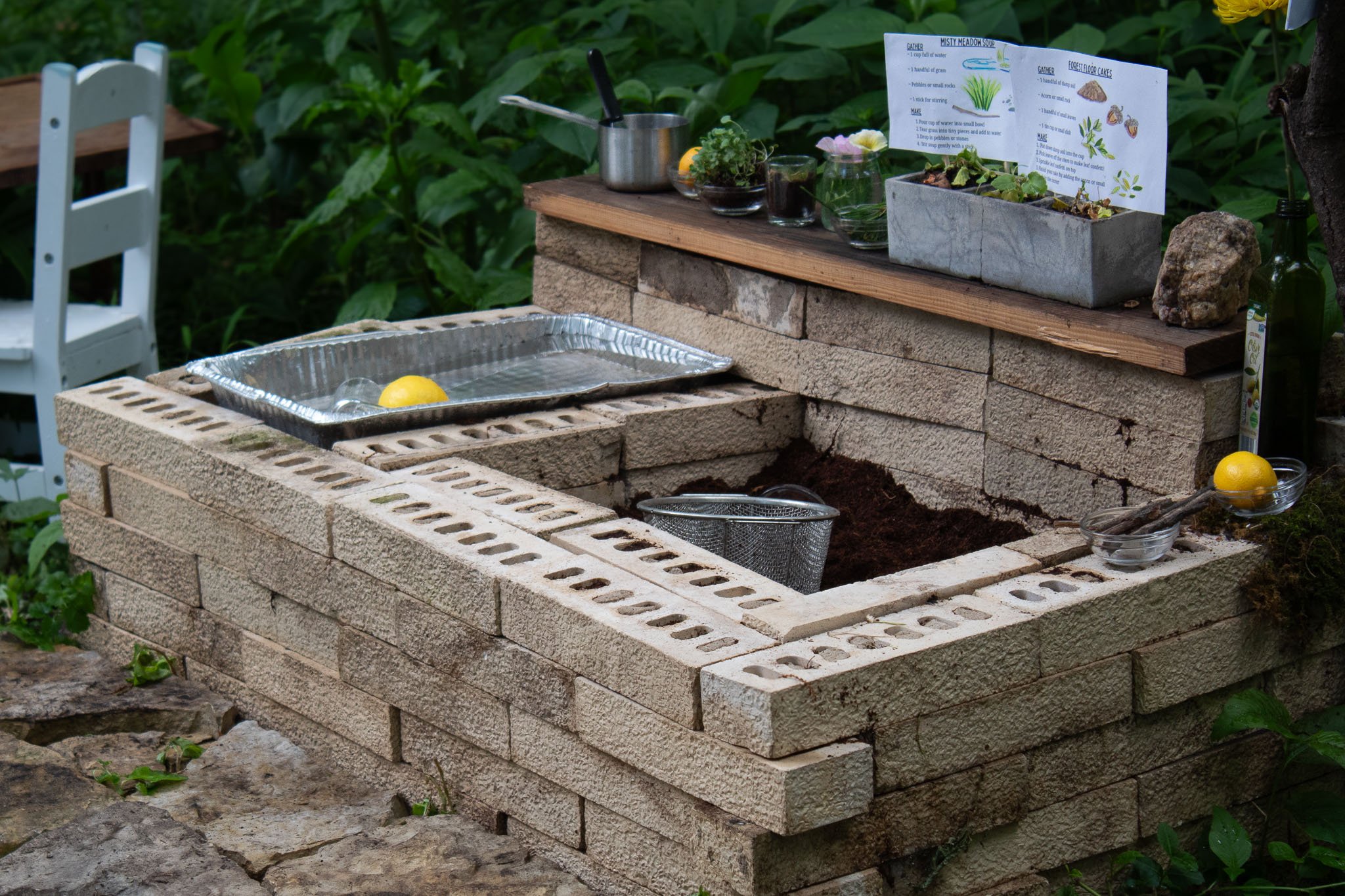 Handmade outdoor mud kitchen built from stacked blocks with a water tray, soil play area, natural ingredients, recipe cards, utensils, and lemons surrounded by garden greenery.