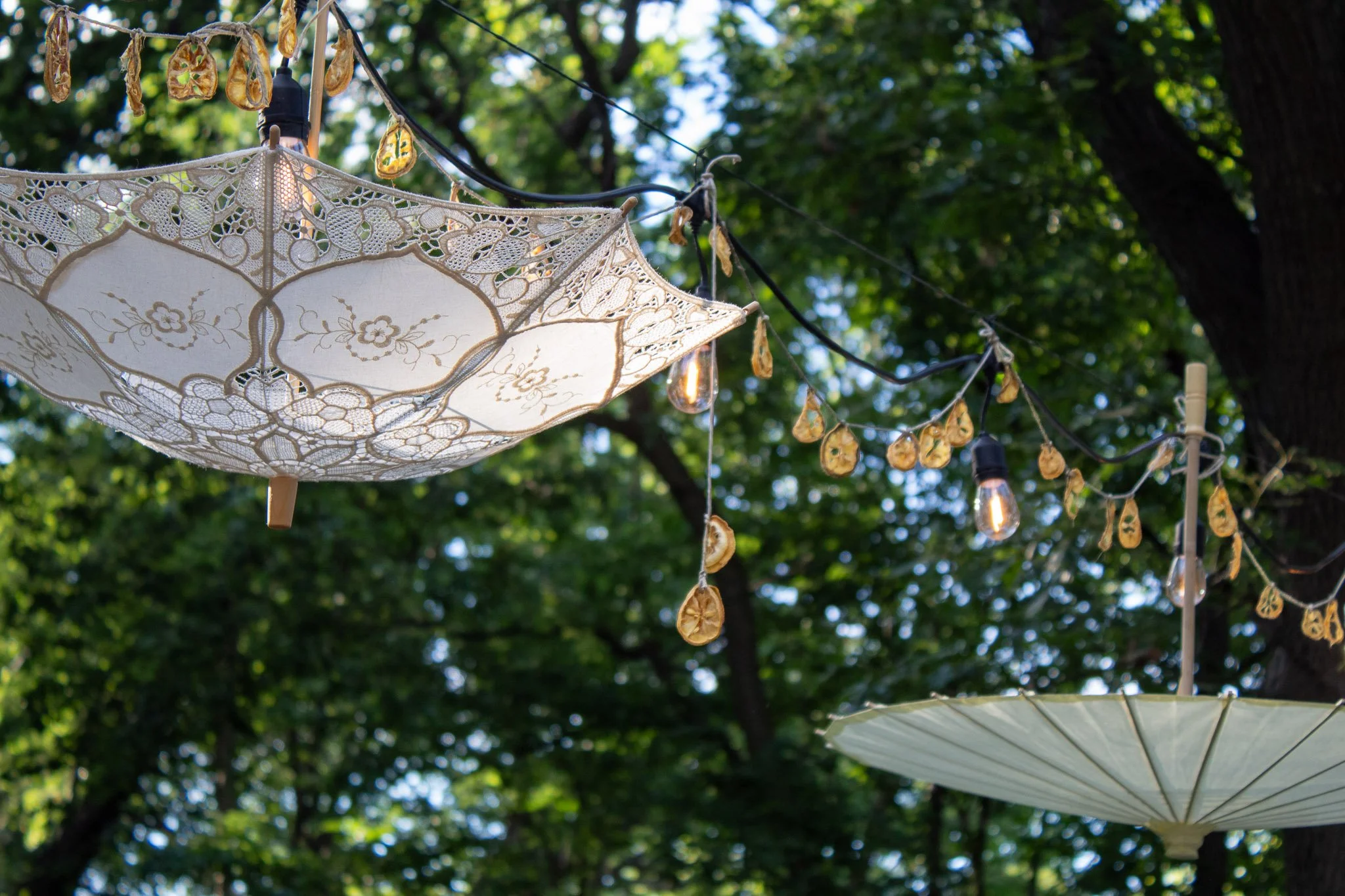 Lace parasols, dried lemon garlands, and glowing string lights suspended beneath the trees at a garden birthday party.