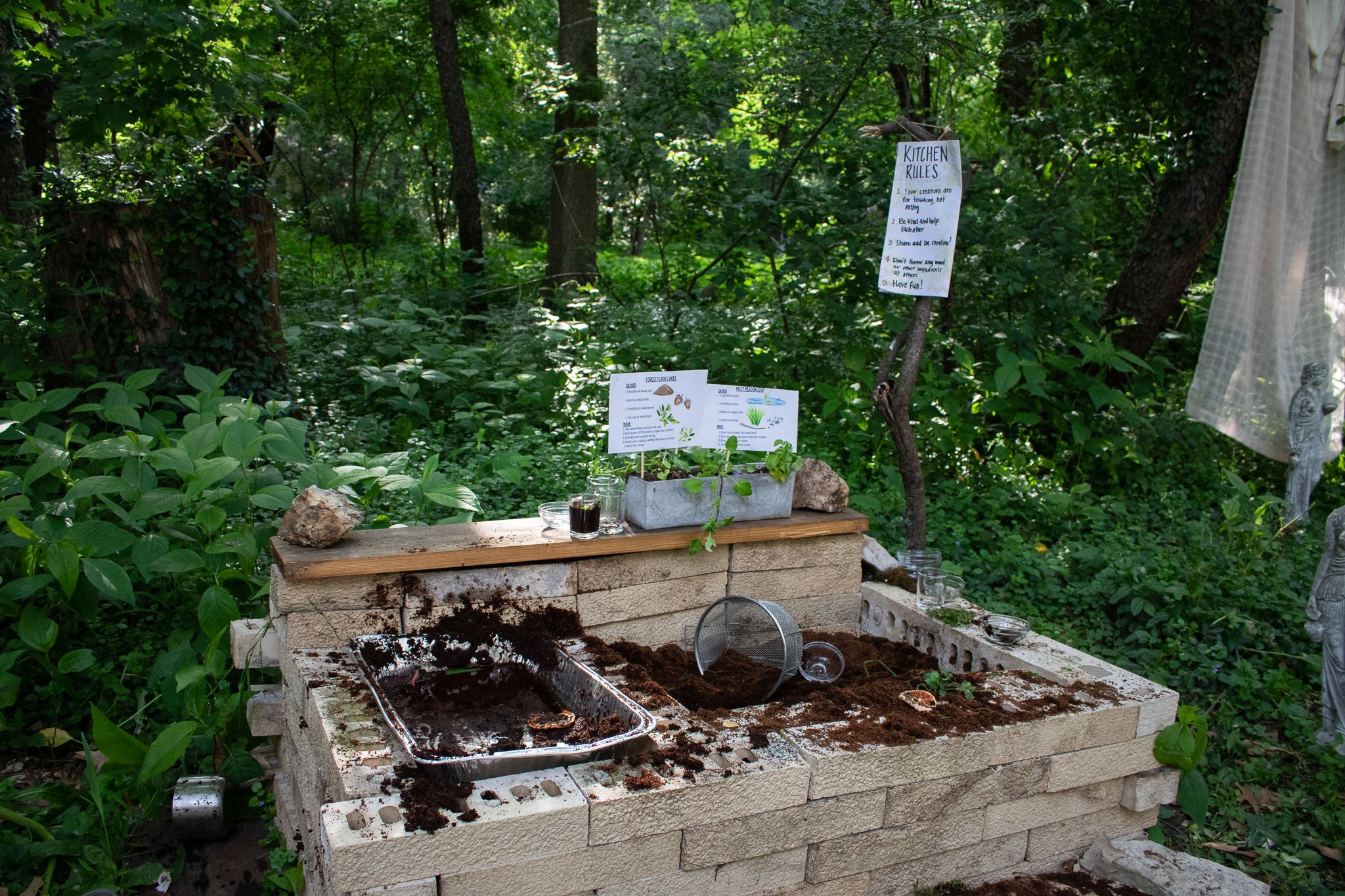 DIY mud kitchen made from bricks, wood, stone with soil, water, cups, and natural materials in a wooded backyard for a garden first birthday party.