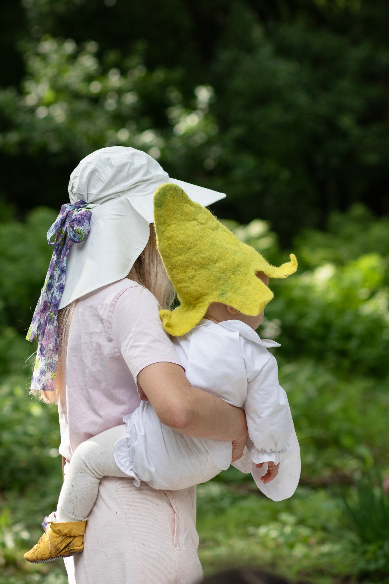Mother holding baby in a handmade yellow Lemon Baby hat, white first birthday outfit, and mustard booties in a garden setting.