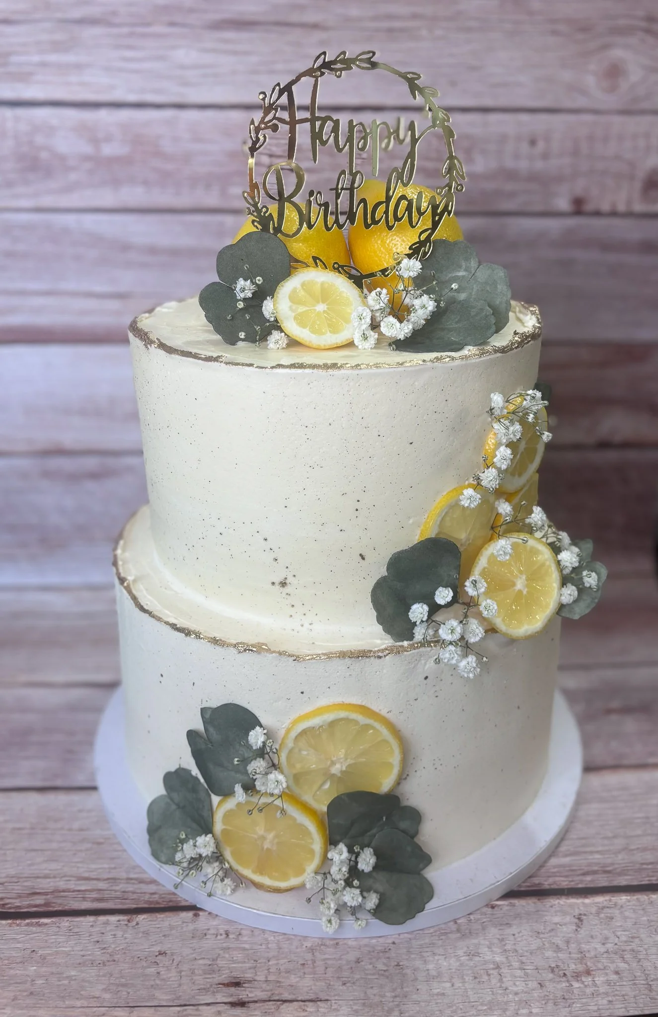 Two-tier vegan strawberry birthday cake decorated with lemon slices, greenery, white flowers, and a “Happy Birthday” topper for a lemon garden first birthday party.