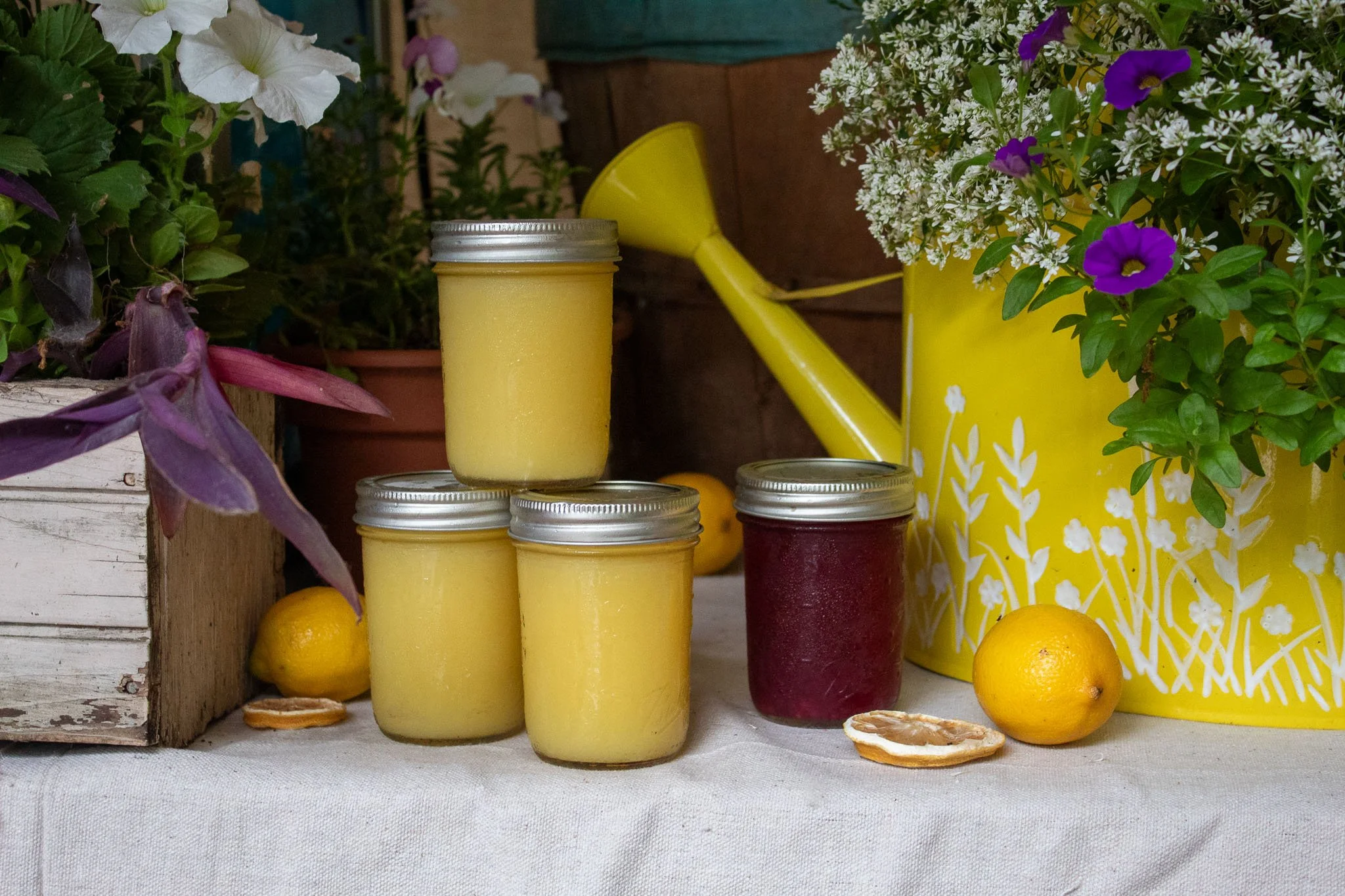 Homemade honey-lemon and berry snow-cone syrups in mason jars beside flowers, lemons, and a yellow watering can.