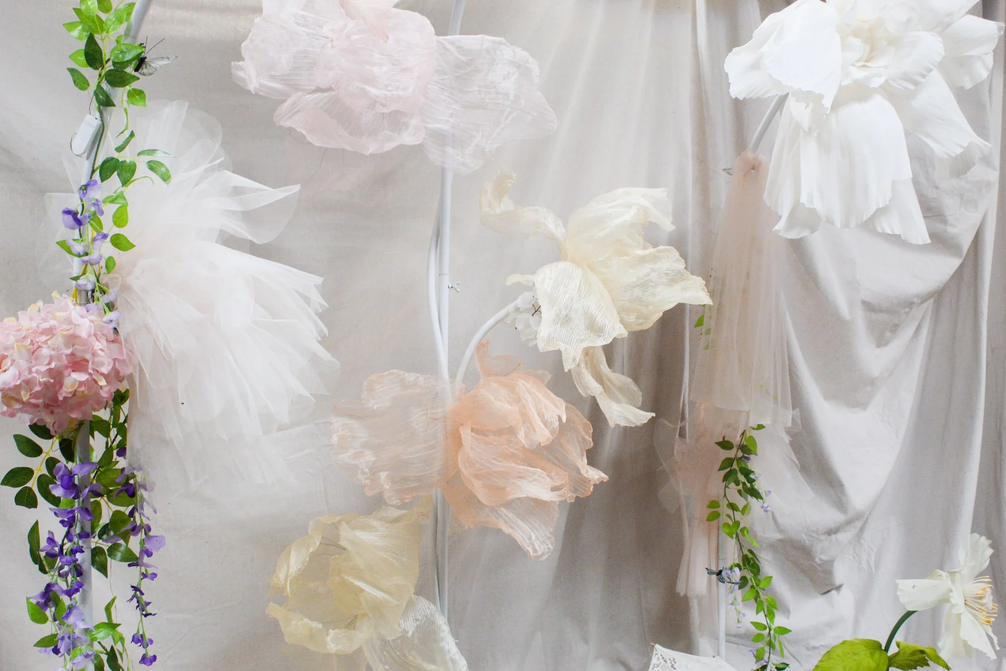 Oversized handmade flowers in white, blush, peach, and pale yellow are arranged against soft draped fabric with trailing green and purple vines for a whimsical garden first birthday party.