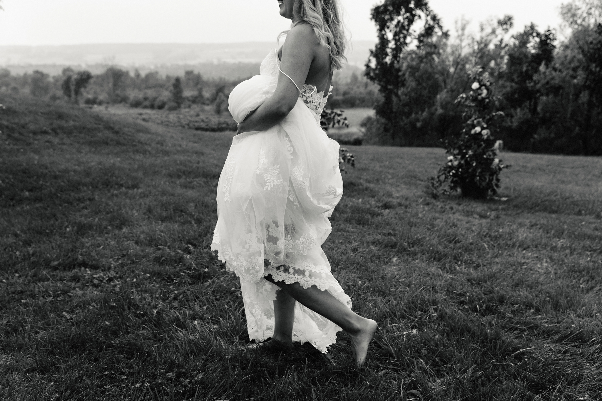 Bride walks barefoot in a field while holding her lace gown at C