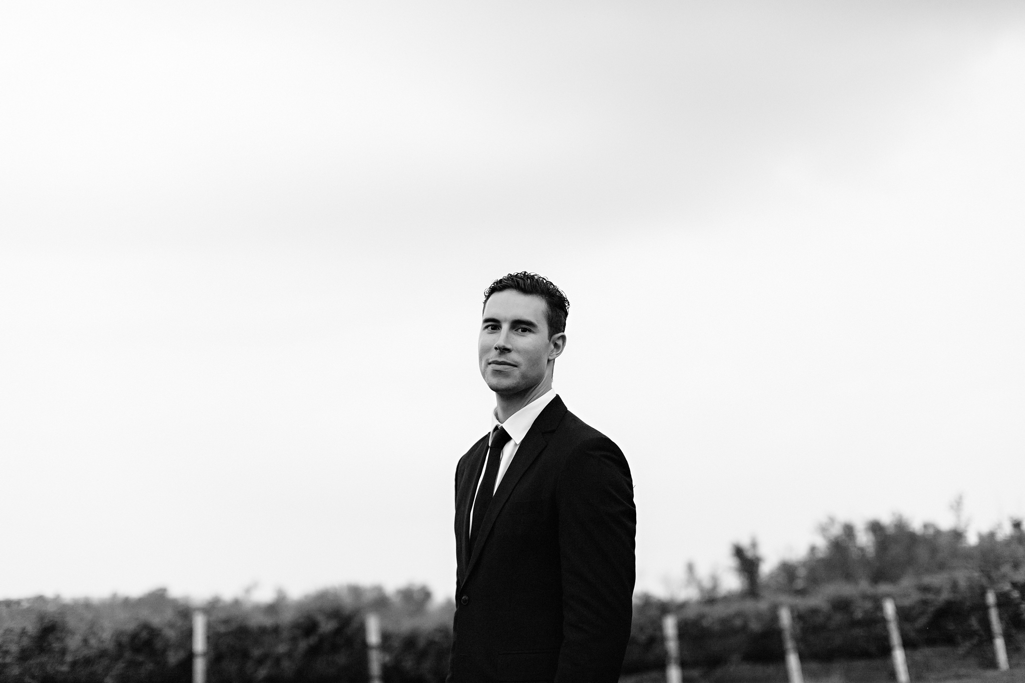 Handsome groom stands in the vineyards in classic black suit at 