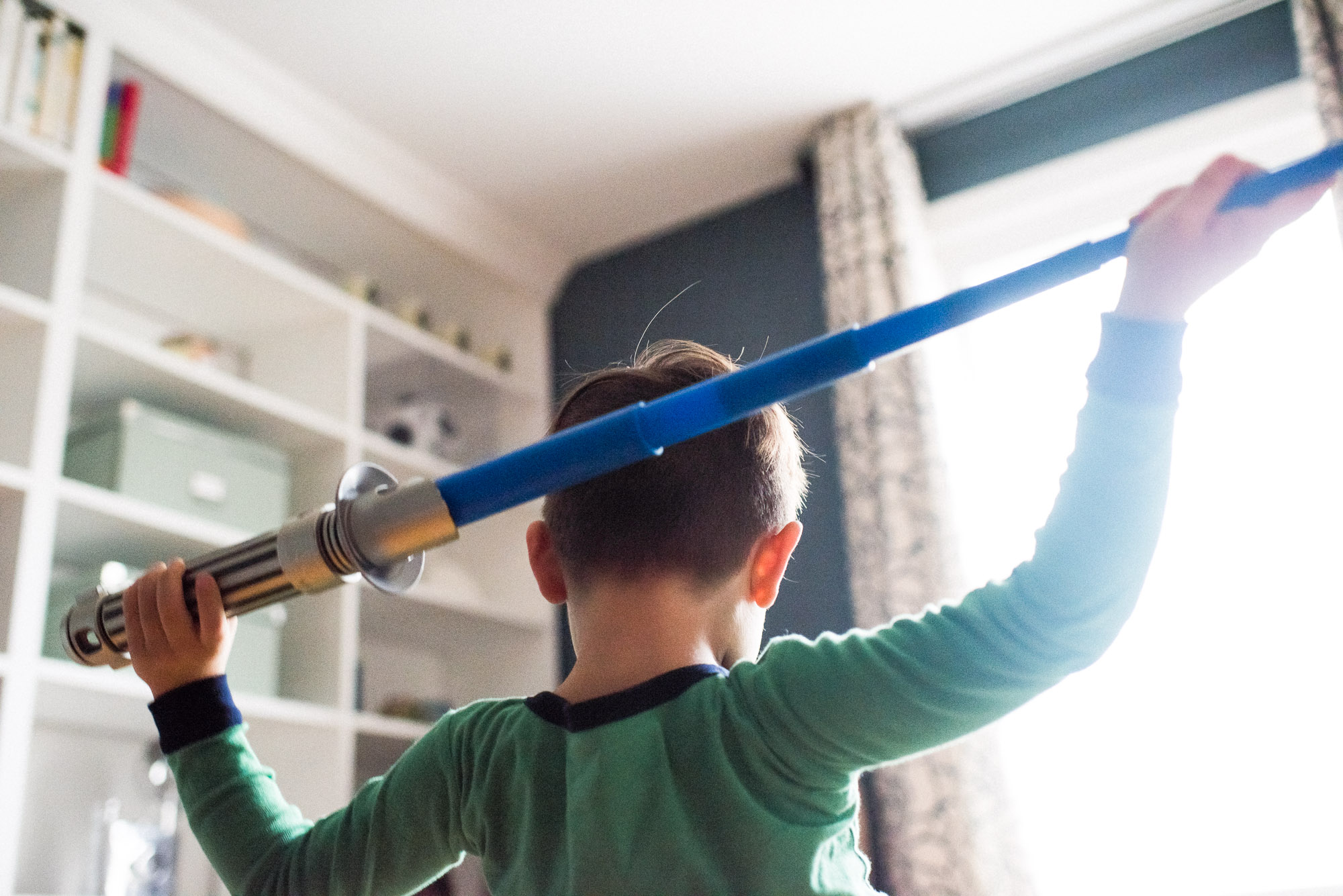 boy holds lightsaber behind his head in front of window