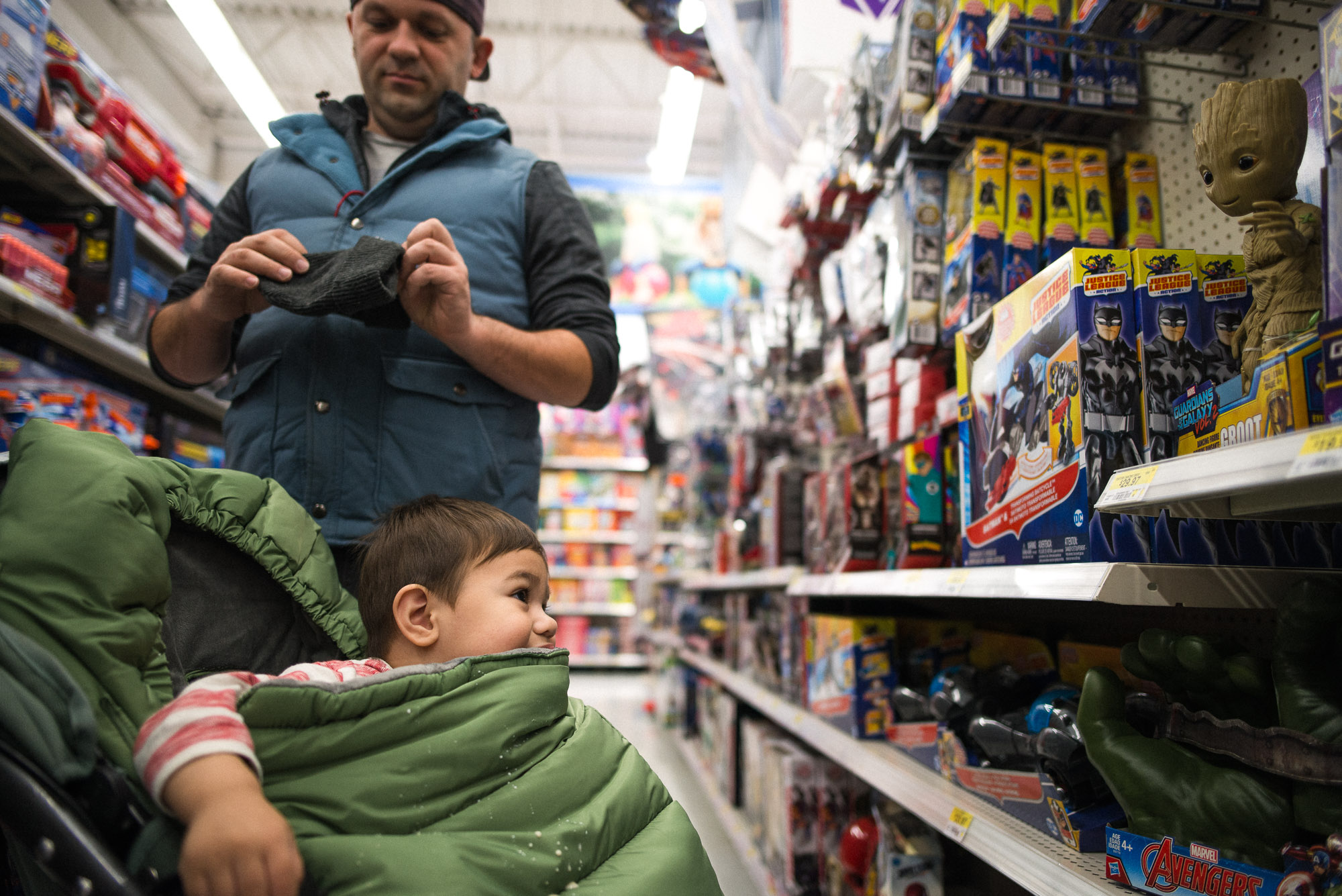 dad distracted while toddler in stroller looks at toys on the shelf