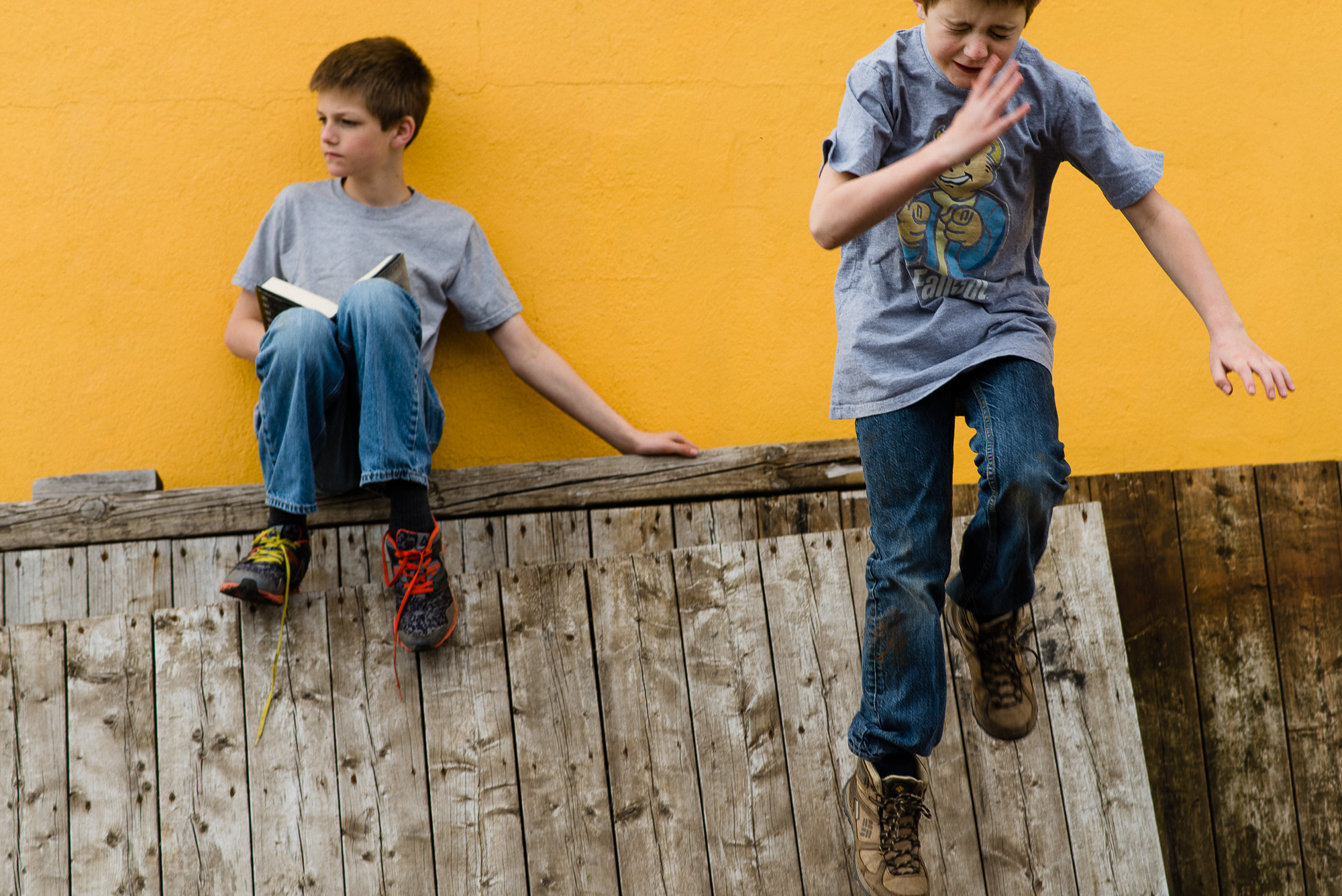 boy jumps from a wooden crate as his brother sits and reads