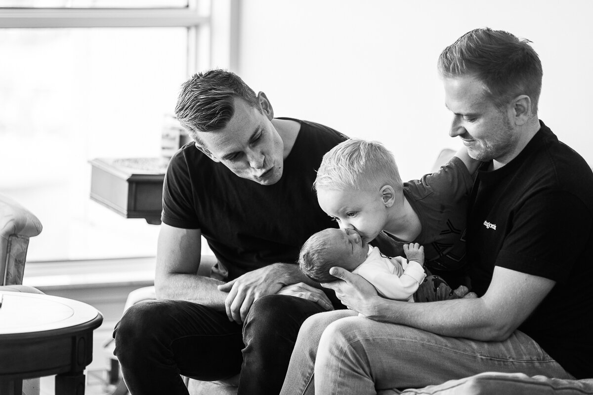 little brother gives his new baby brother a kiss while being held by their dads 