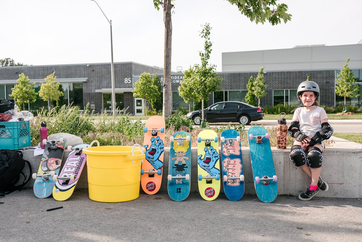 Girl wearing grey helmet is sitting on the bench with a lot of skateboards nearby.