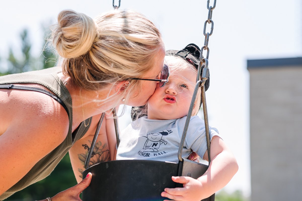 Mother kissing baby  when he is sitting on the swing.