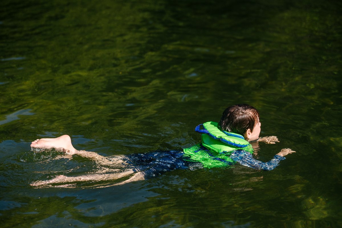 Boy swimming in the water wearing green coloured safety jacket/].