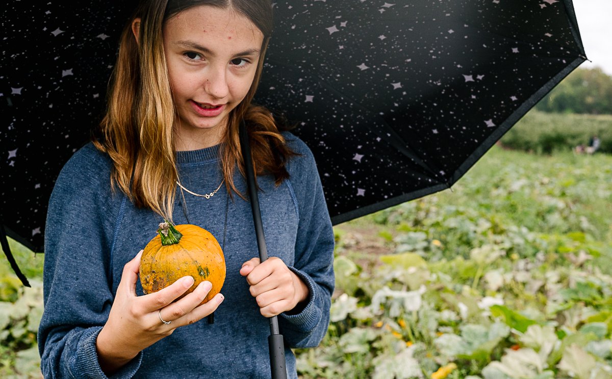 Girl holding yellow pumpkin in one hand and black umbrella in another.