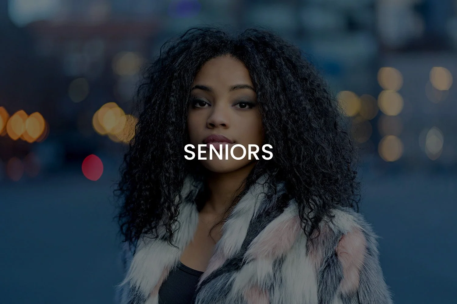 A young woman with curly black hair standing outdoors during twilight, wearing a fur coat, with the word 'SENIORS' written across her chest.