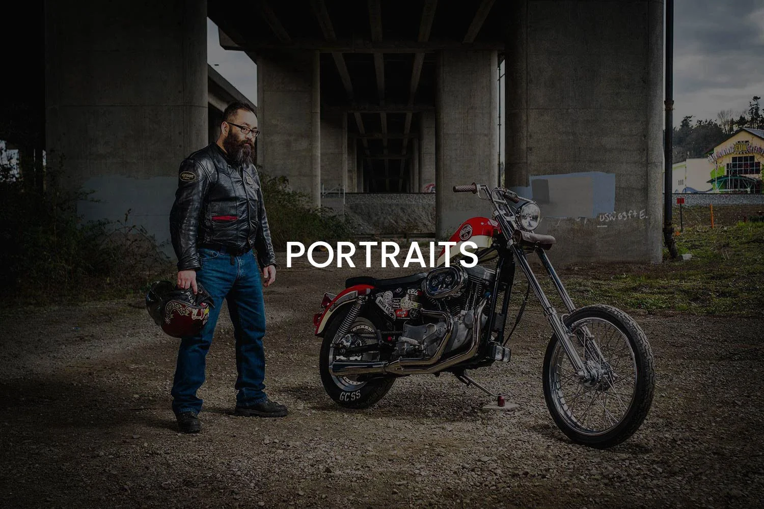 A bearded man in a leather jacket holding a helmet stands under a bridge near a vintage motorcycle with a underpass and graffiti in the background.