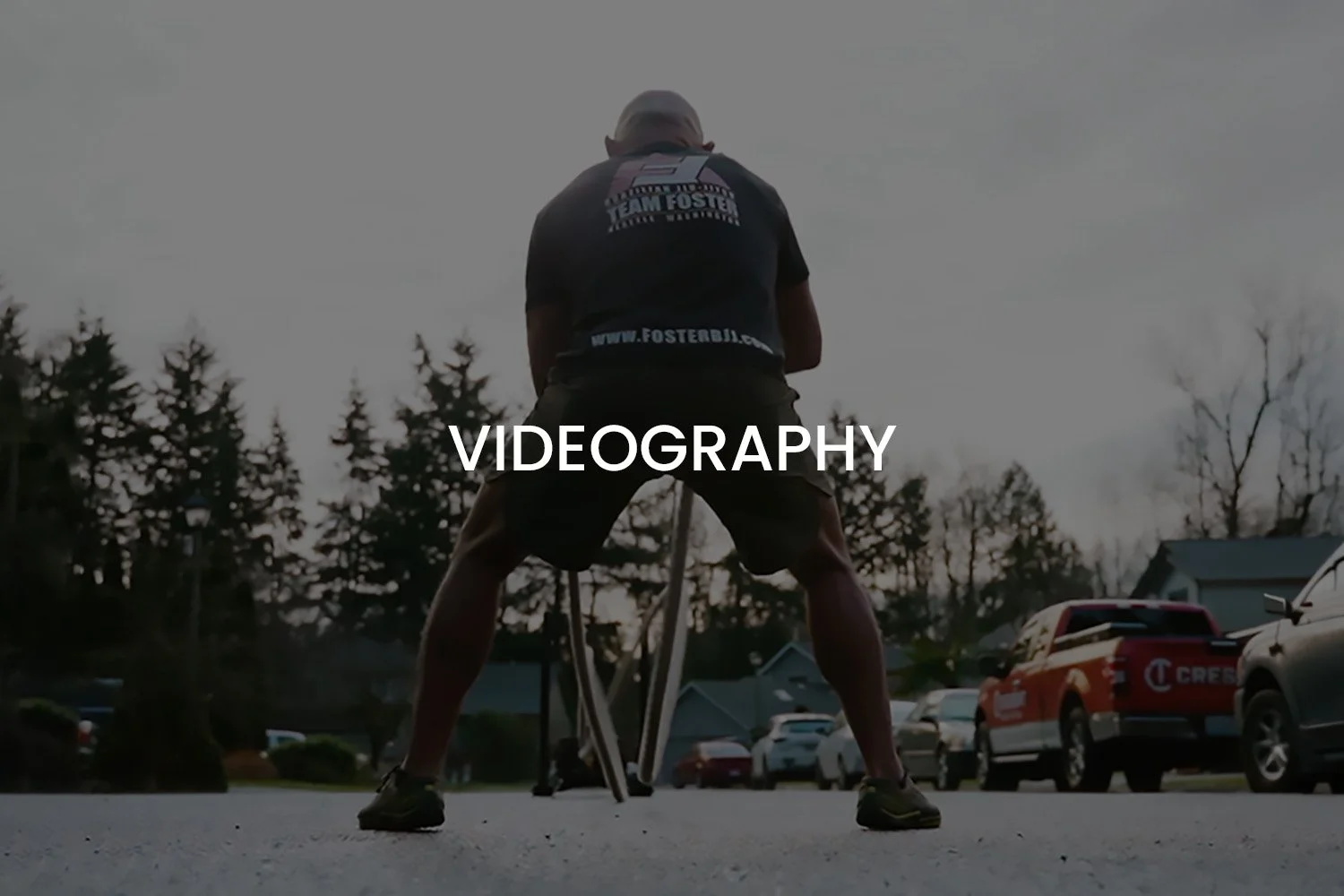 A person is in a squatting position outdoors, ready to start a race, with a tripod or crutch-like equipment in front of them, during twilight or early evening. The background shows trees, parked cars, and a cloudy sky.
