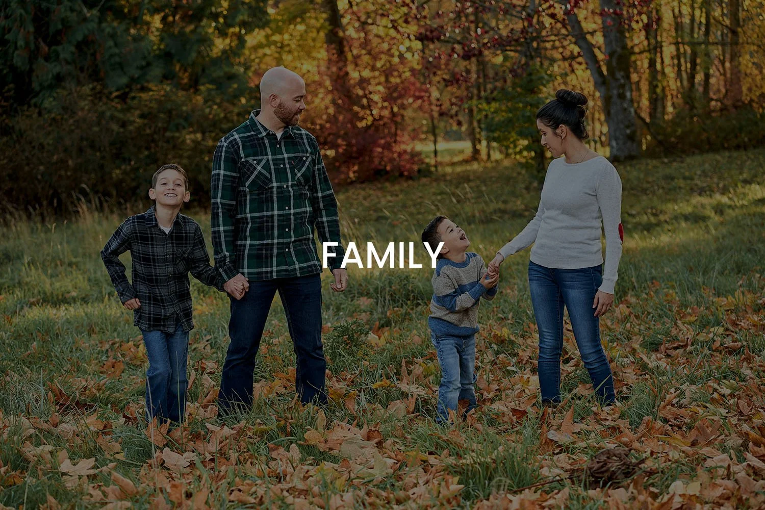 A family of five, including two adults and three children, enjoying a walk in a park during fall. They are holding hands and smiling, with autumn leaves on the ground and trees with colorful foliage in the background.