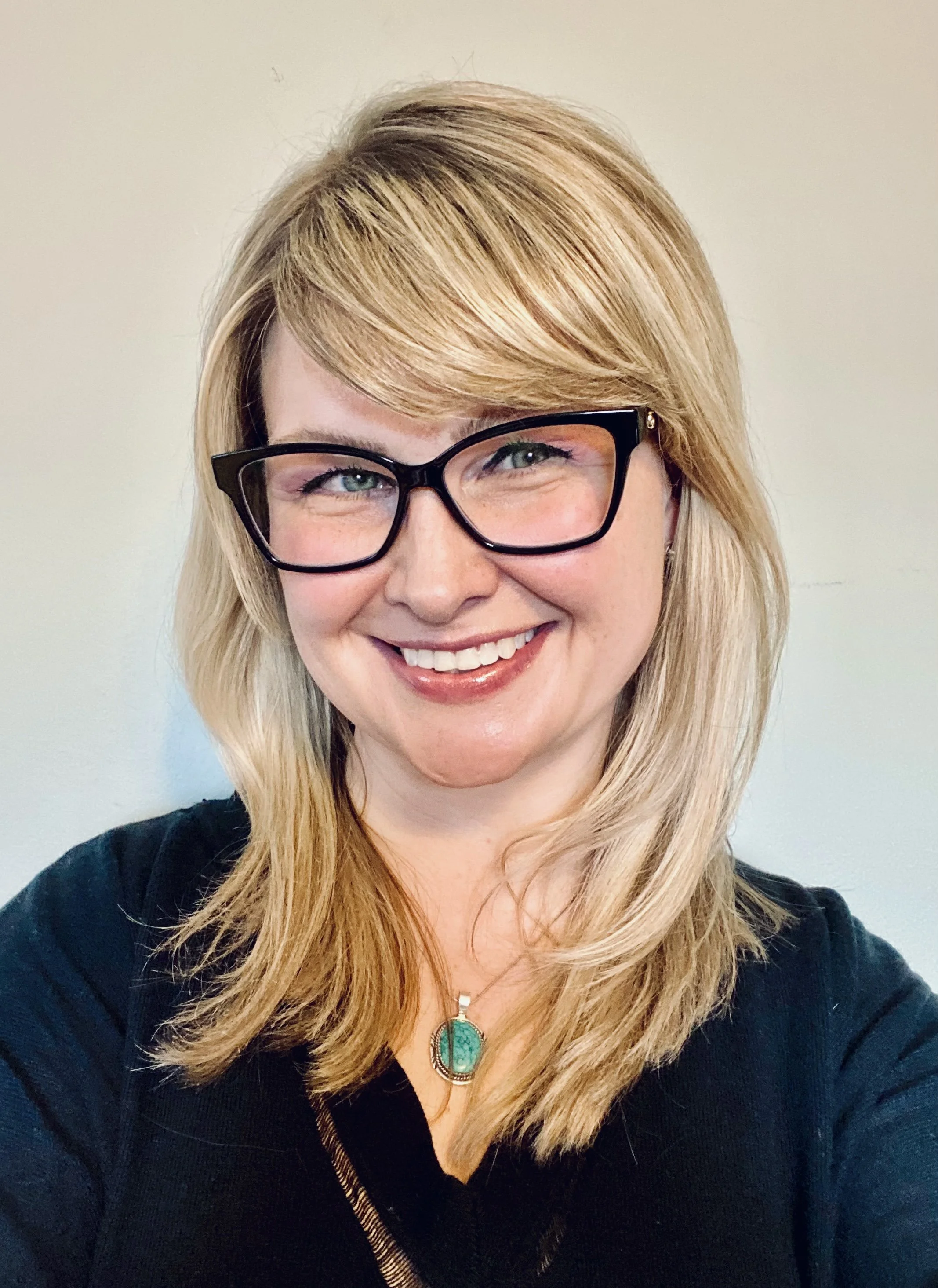Close-up of a smiling woman with blonde hair, wearing black glasses and a turquoise pendant necklace. She is dressed in black and standing against a light-colored background.