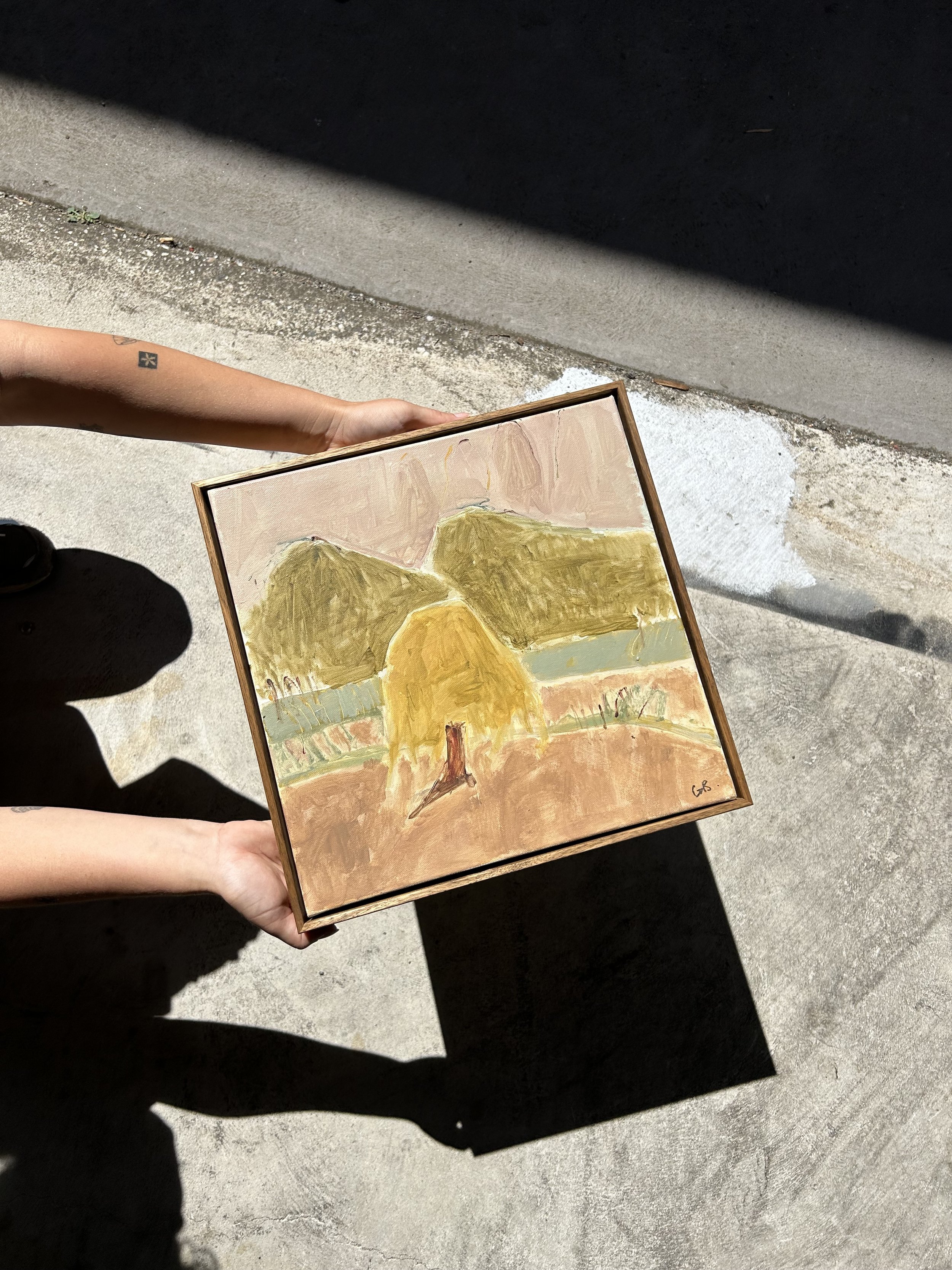 Person holding a framed painting. The background includes muted, abstract colors and a metal shutter.