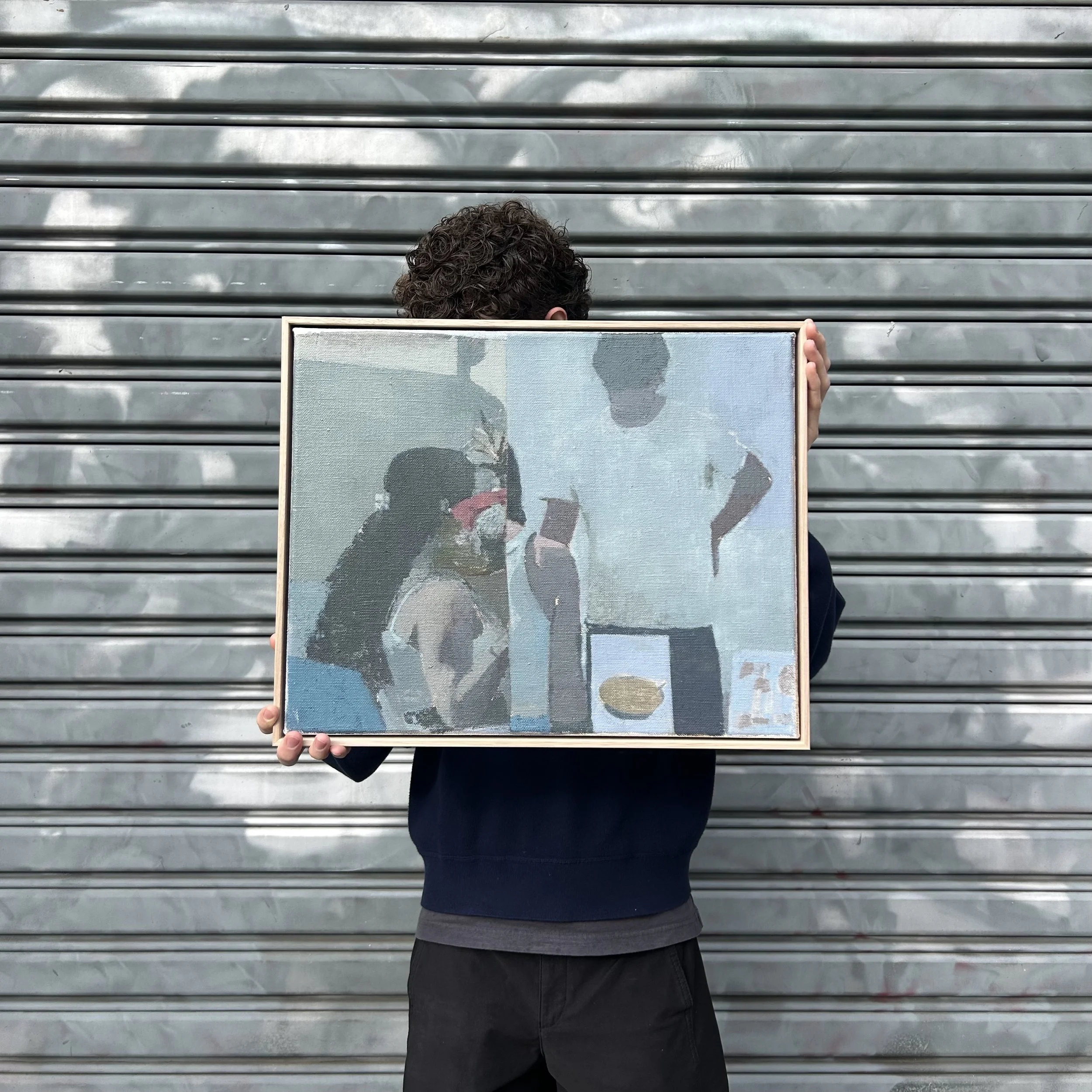 Person holding a framed painting. The background includes muted, abstract colors and a metal shutter.
