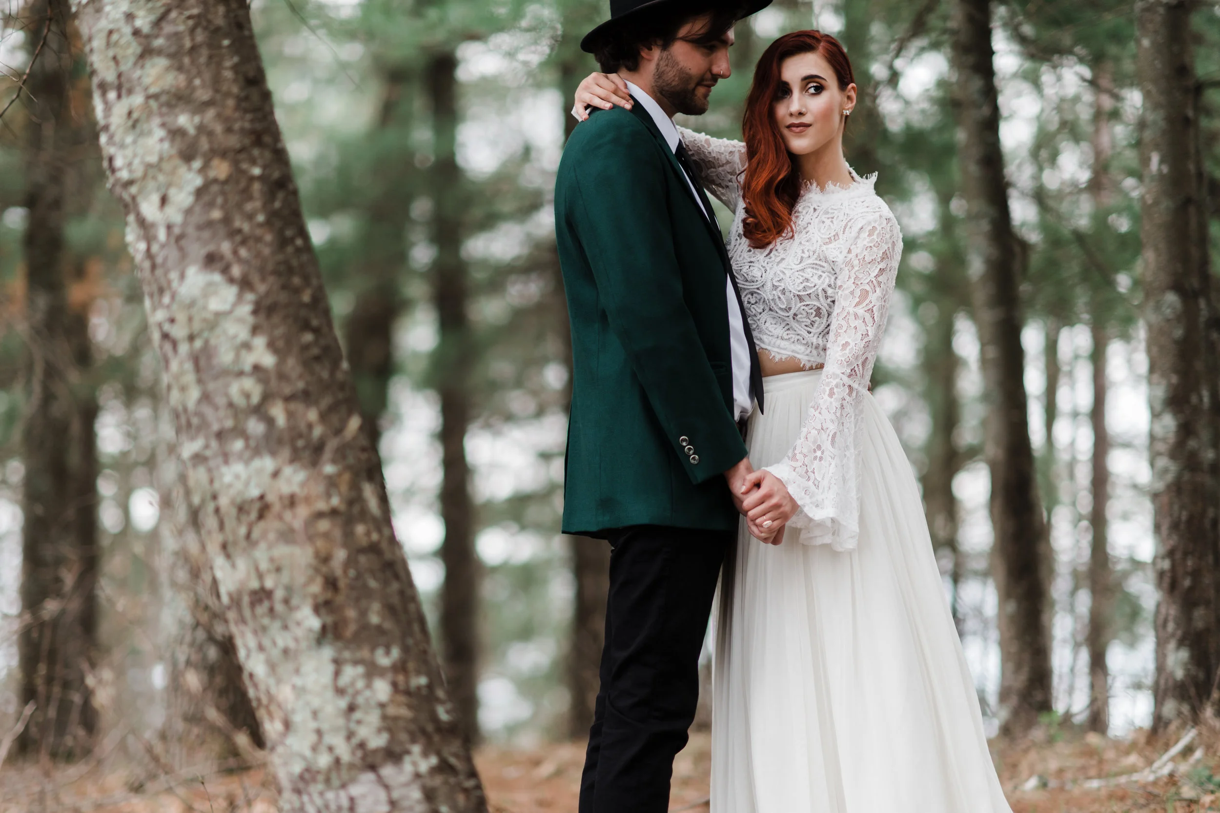 Groom in green jacket and black fedora slow dancing in the woods with bride in lace and beaded crop top and flowing skirt.
