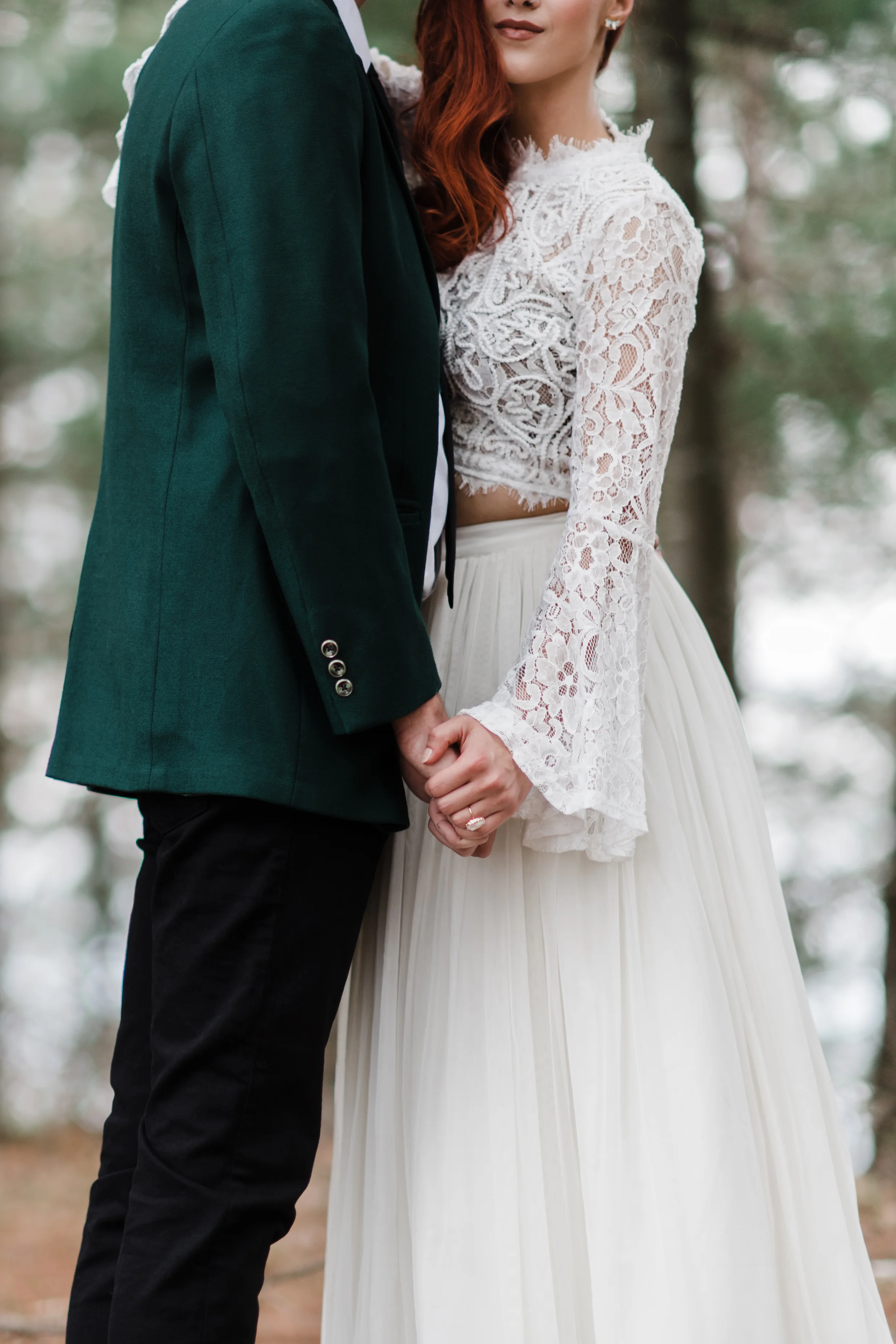 Groom in green jacket and black fedora slow dancing in the woods with bride in lace and beaded crop top and flowing skirt.