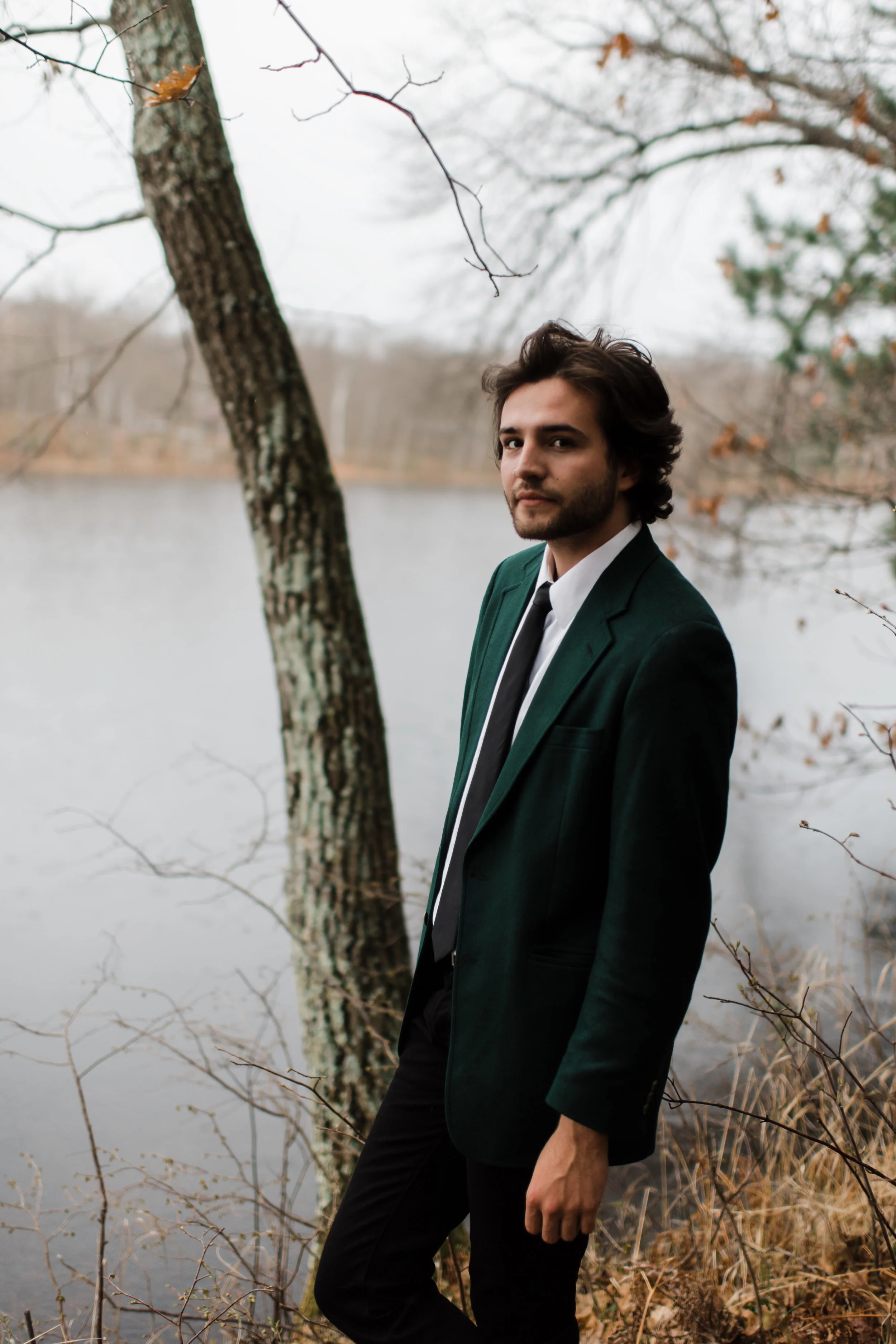 Groom in green jacket and black fedora stands casually in front of the water.
