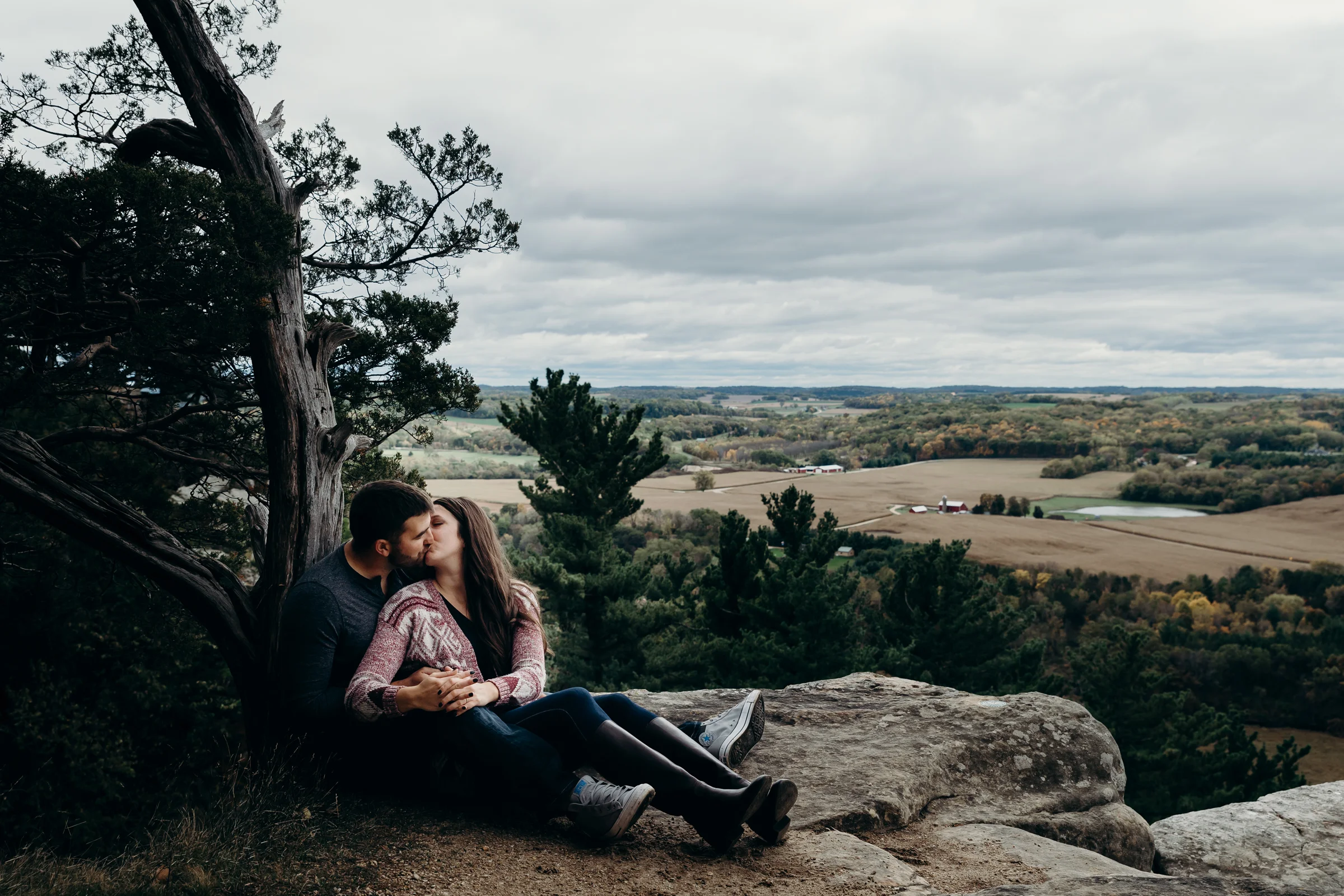 Engaged couple sits and kisses on top of a rock at Gibraltar Rock State Natural Area in Lodi, WI.