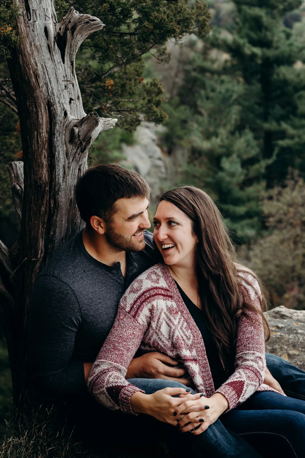 Engaged couple sits back against a tree and shares a laugh.
