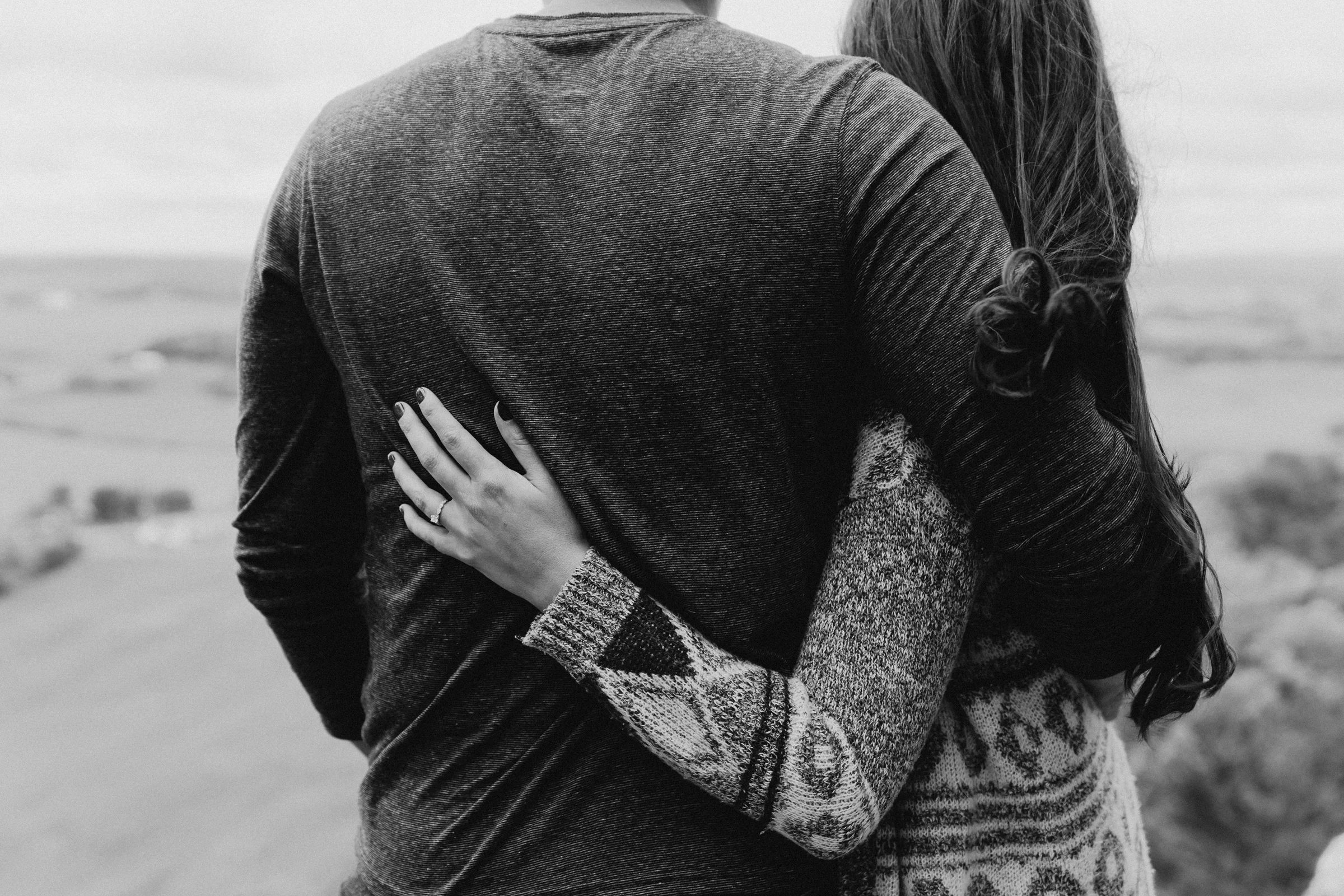 Woman rests her hand on her fiancé's back and shows off her princess cut engagement ring.