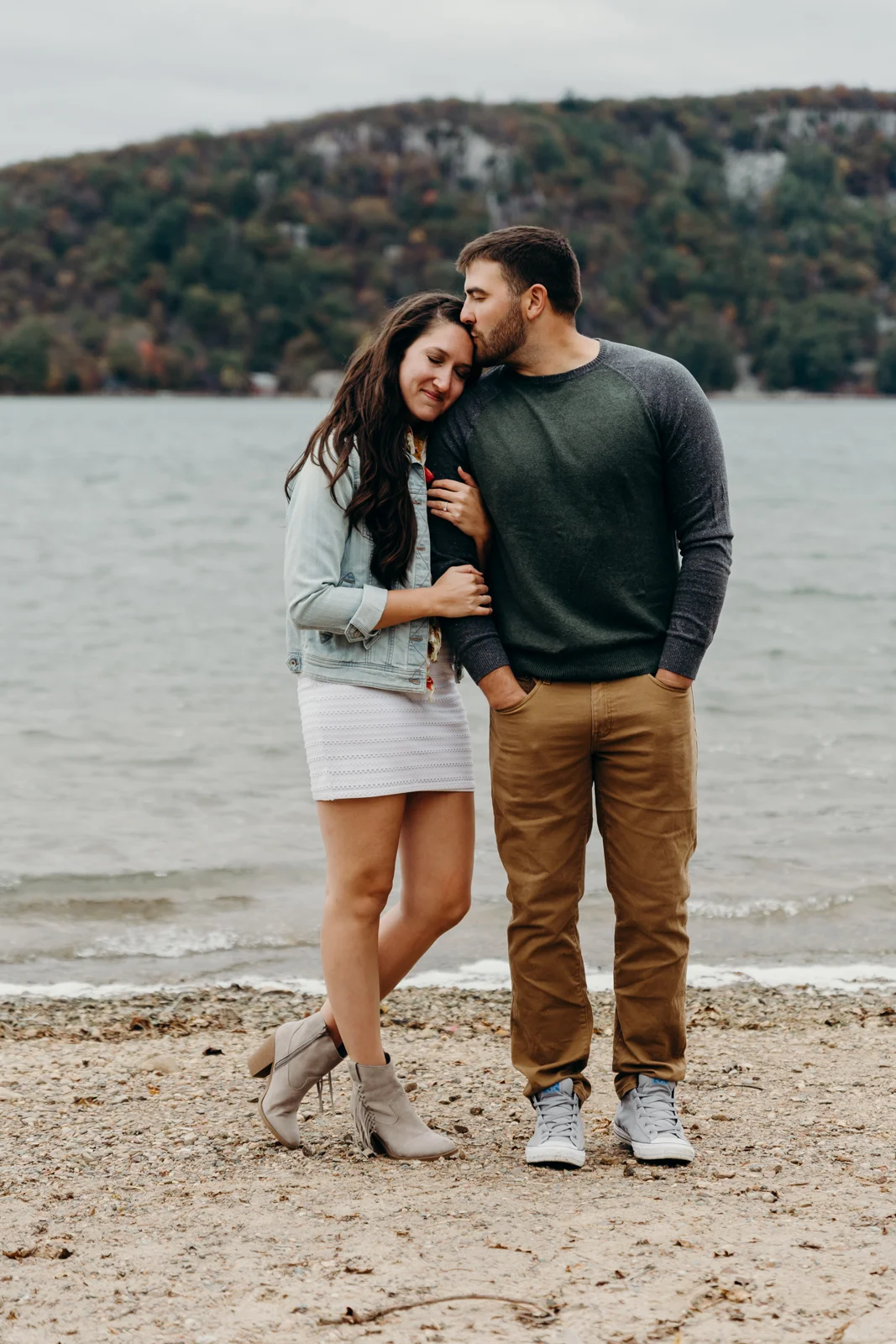 Woman stands next to her fiancé and rests her head on his shoulder as he kisses her hair.