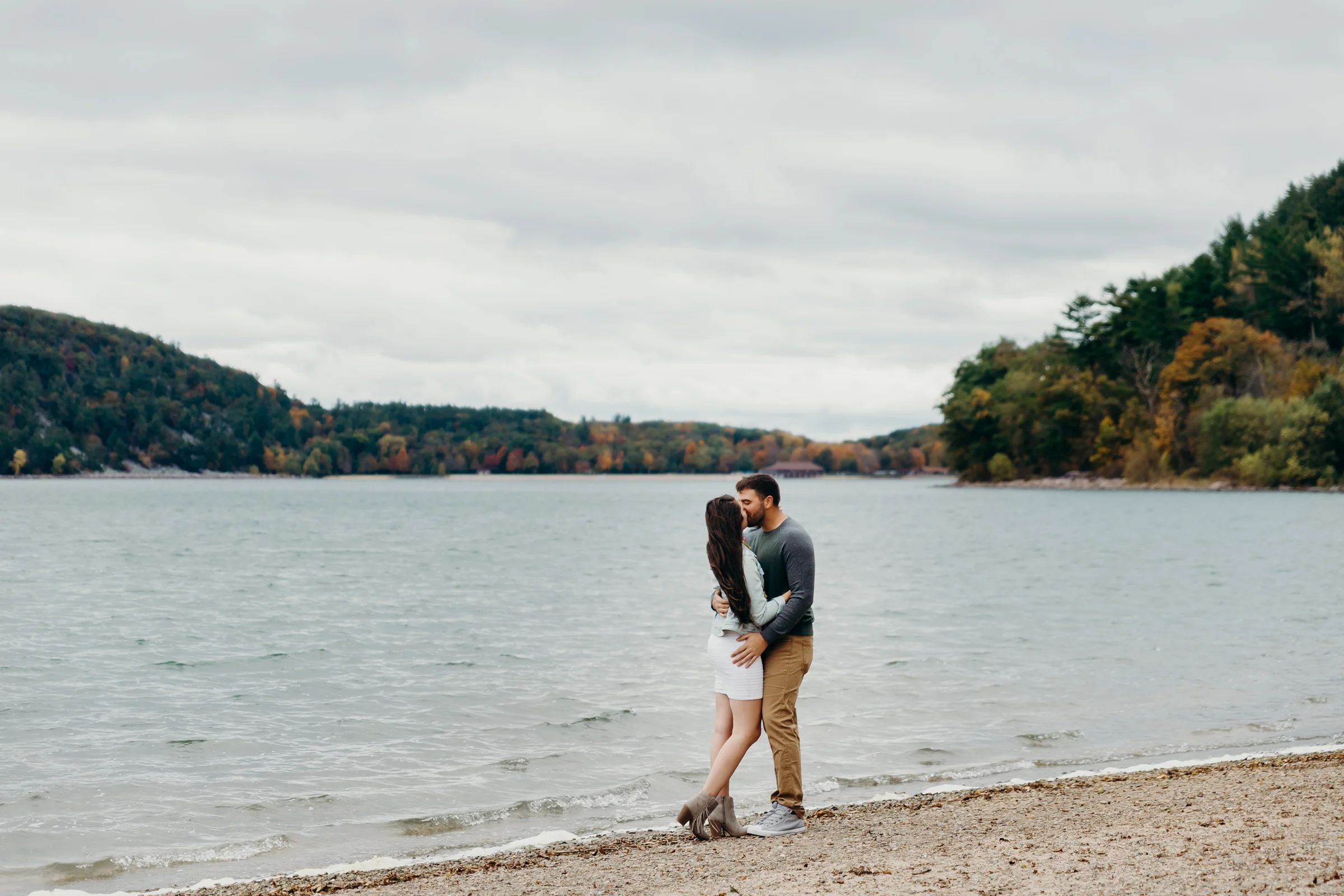 Engaged couple kisses on the beach with beautiful fall colors in the background.