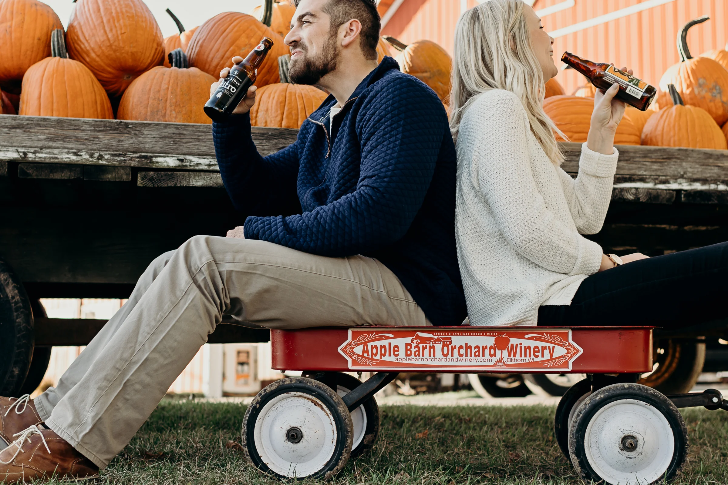 Engaged couple shares a beer while sitting in a red wagon at Apple Barn Orchard and Winery in Elkhart, WI.