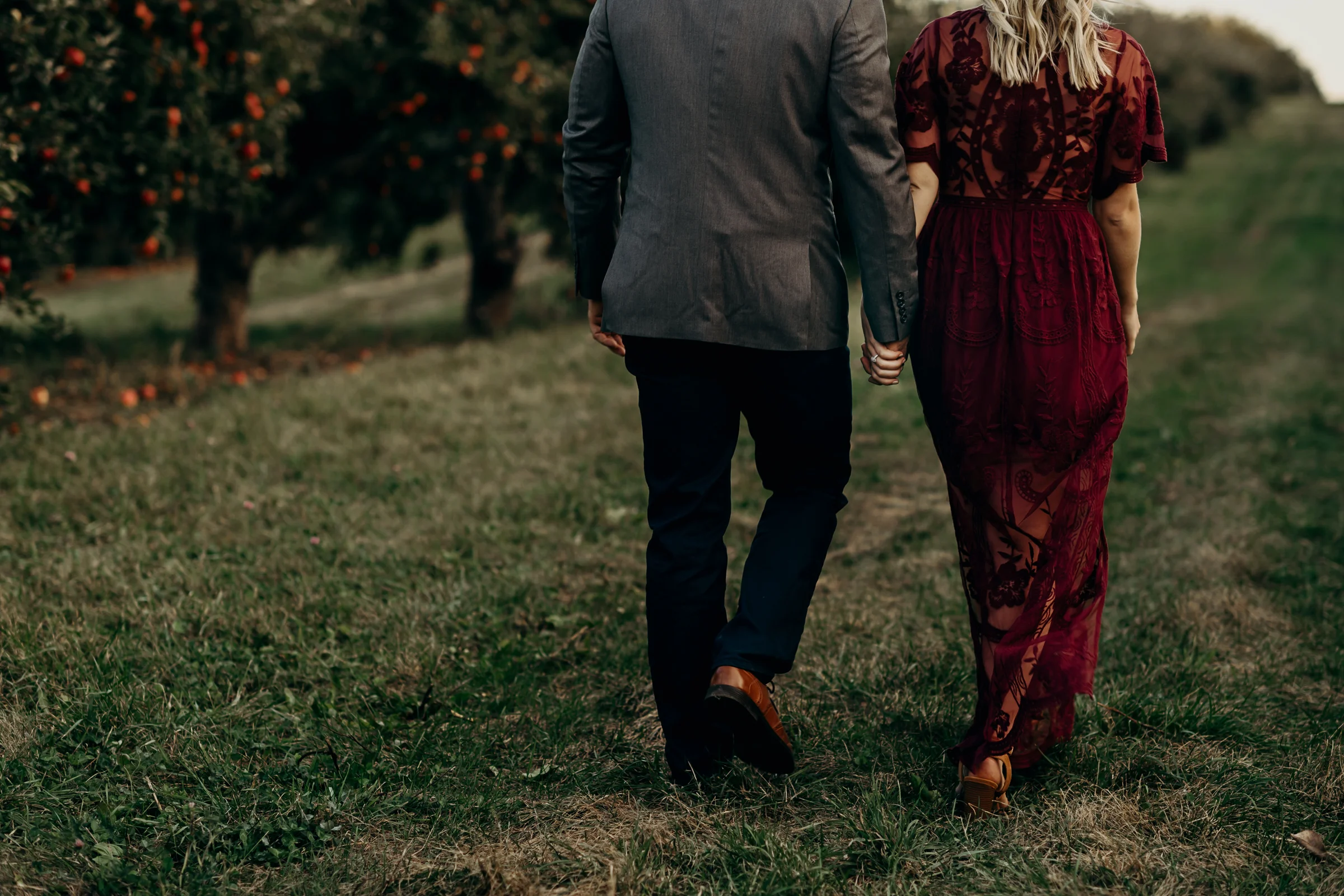 Engaged couple holds hands as they walk through an apple orchard.