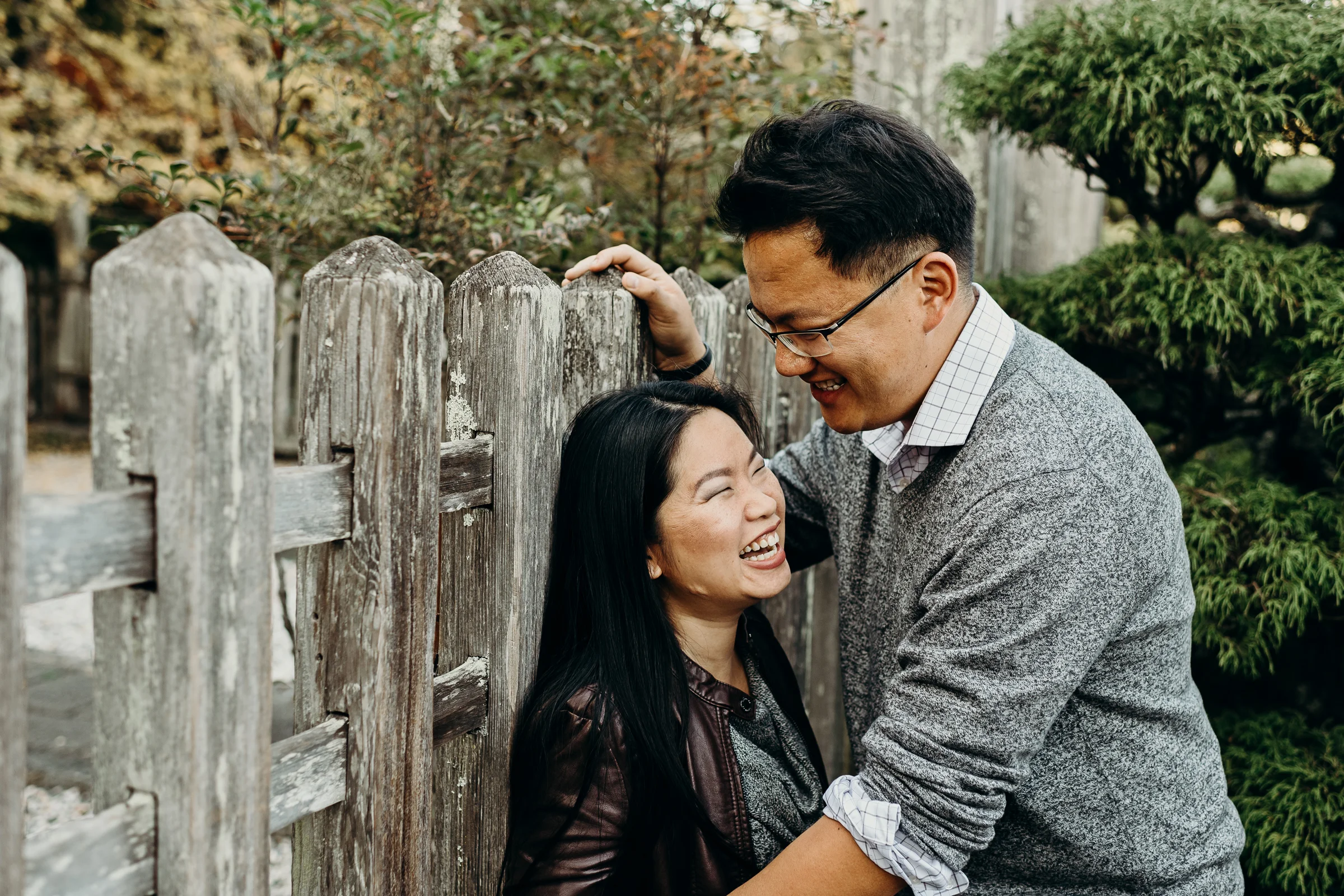 Woman leans back against a wooden fence and laughs as her fiance leans into her.