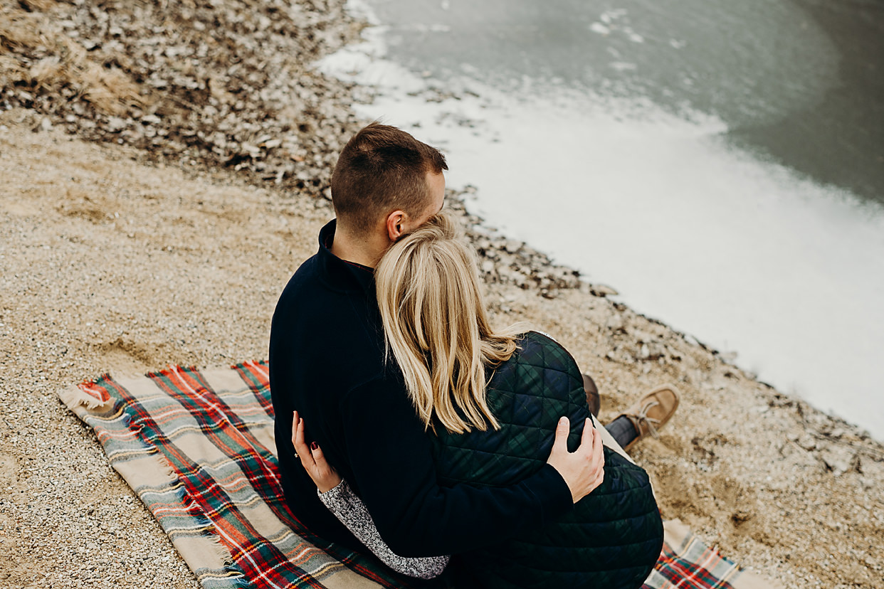 Engaged couple sit and snuggle outside on a blanket