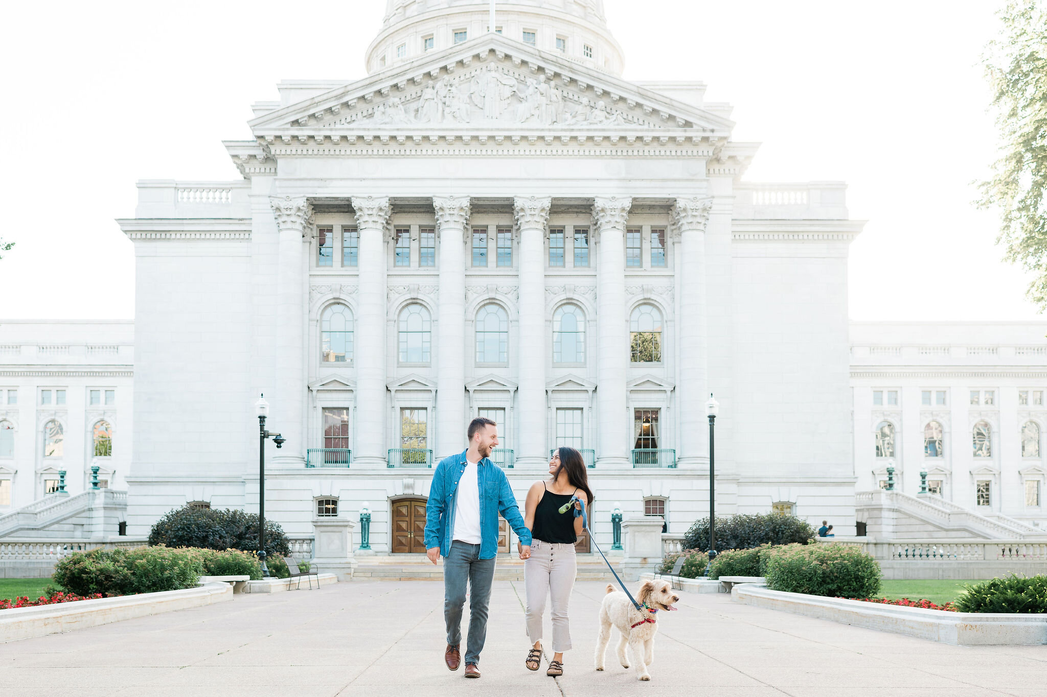 Engaged couple walking with their dog in front of the Wisconsin State Capitol in Madison, WI