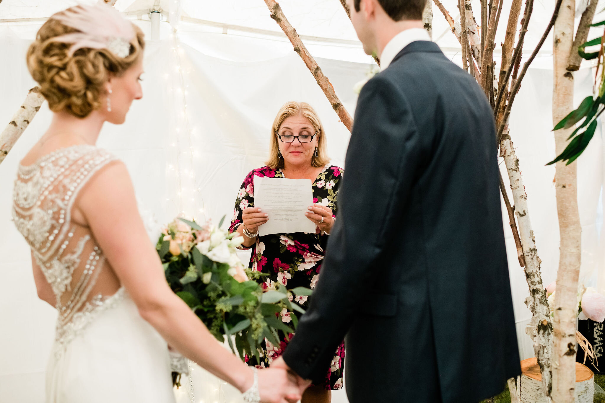 Bride and groom holding hands while wedding guest reads