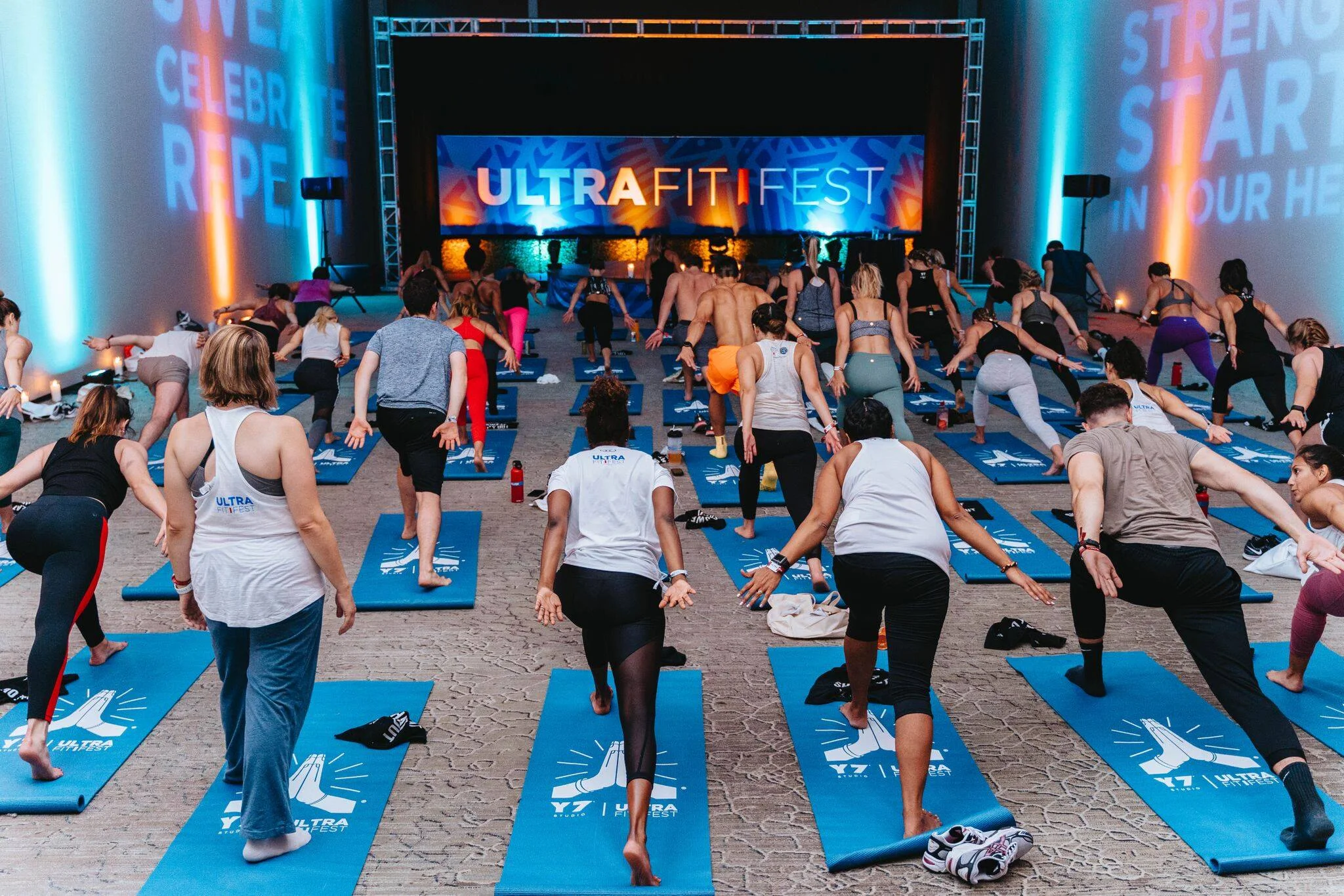 Group of people participating in a yoga or fitness class on blue mats at UltraFit Fest, with a large illuminated sign in the background reading UltraFit Fest and colorful lighting on the walls.