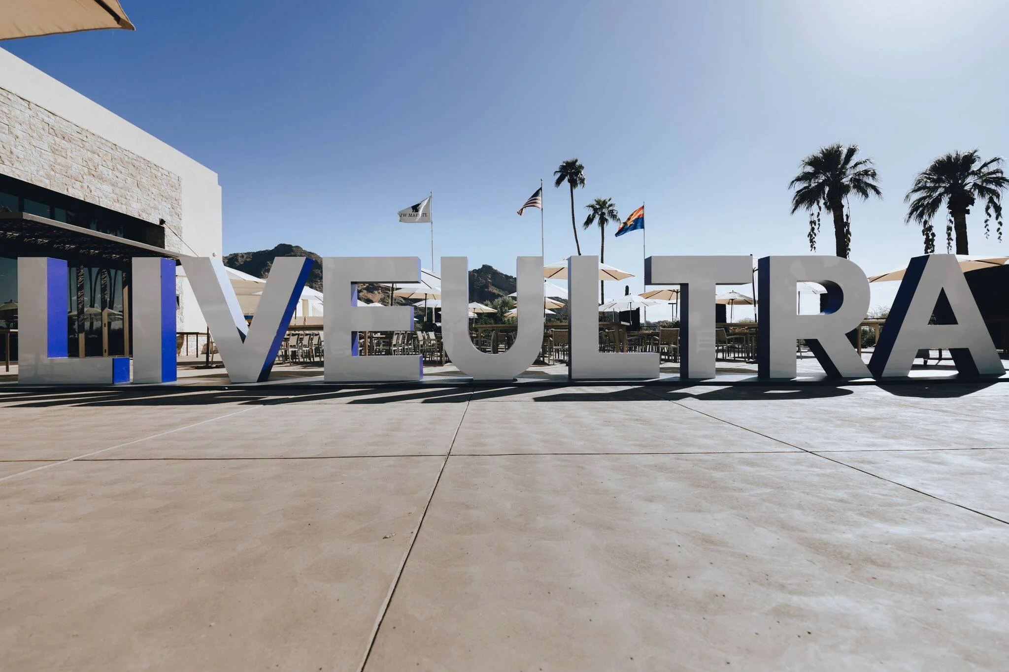 Large white letters spelling 'VUE' on a patio, with beach chairs and umbrellas, palm trees, flags, mountains, and clear blue sky in the background.