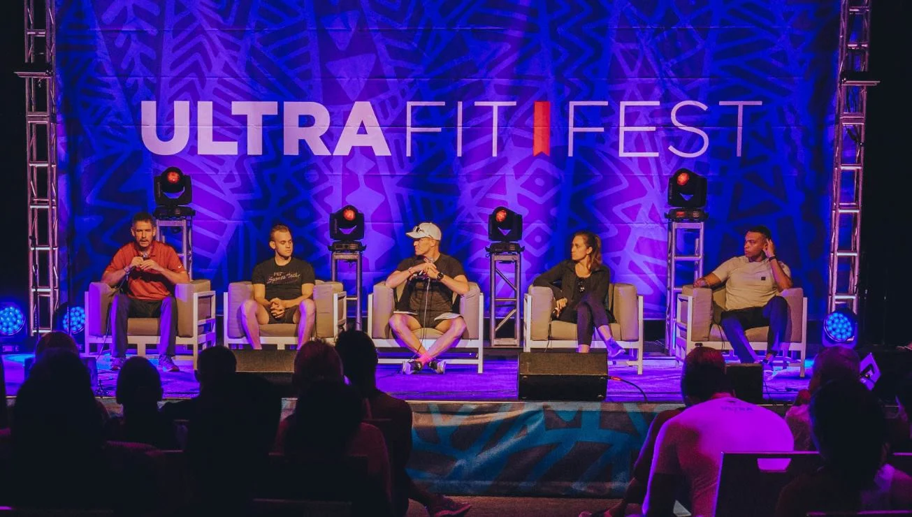Panel discussion at Ultra Fit Fest with five people seated on stage, purple and blue lighting, and an audience watching.