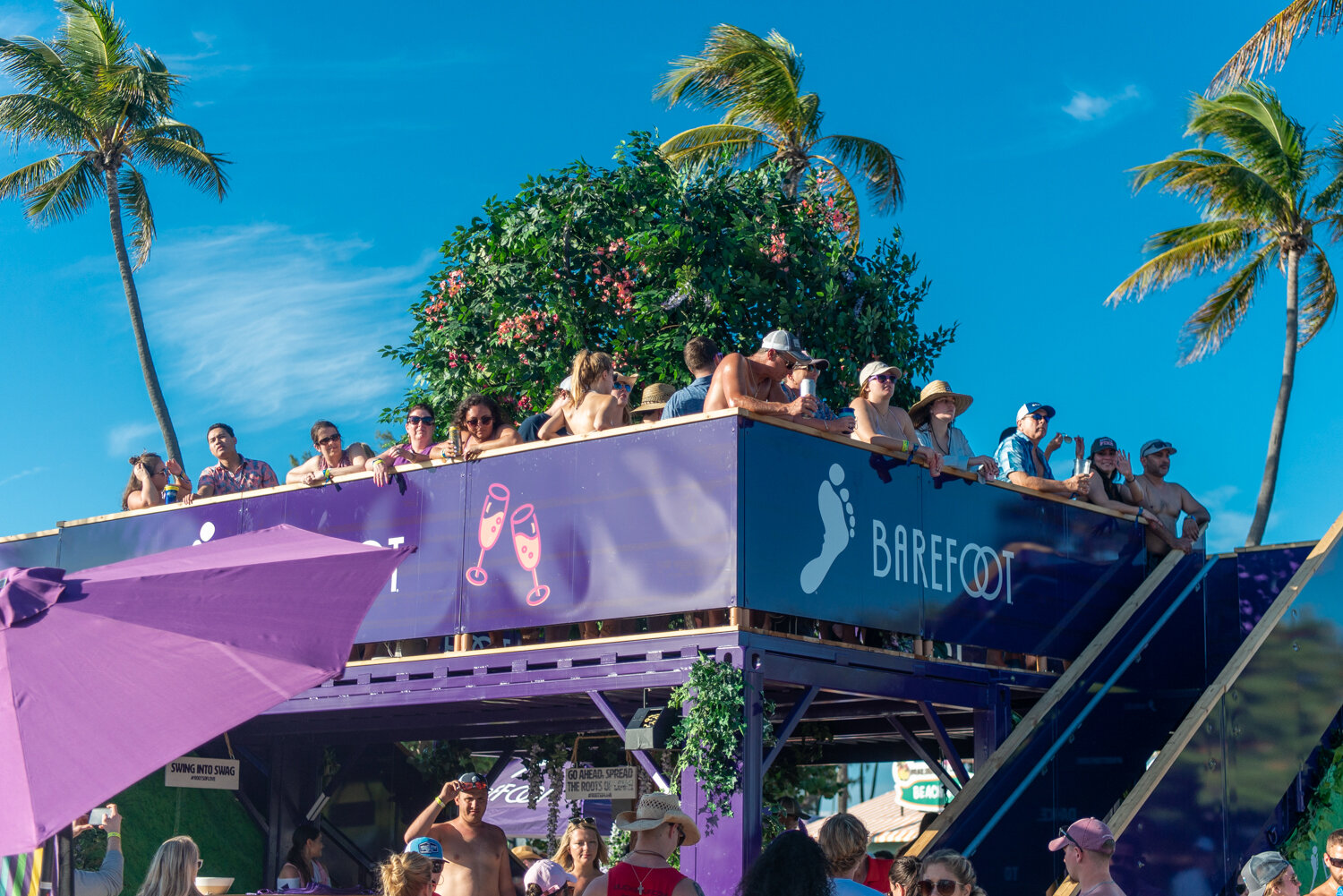 People enjoying a daytime outdoor event on a two-story purple float with palm trees and bright blue sky in the background. The float has a sign that says "BAREFOOT" and images of champagne glasses.