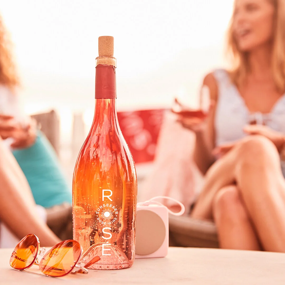 A pink bottle of Rosé wine with sunglasses and a speaker in the foreground, and two women sitting on a patio in the background, one woman smiling and holding a drink.