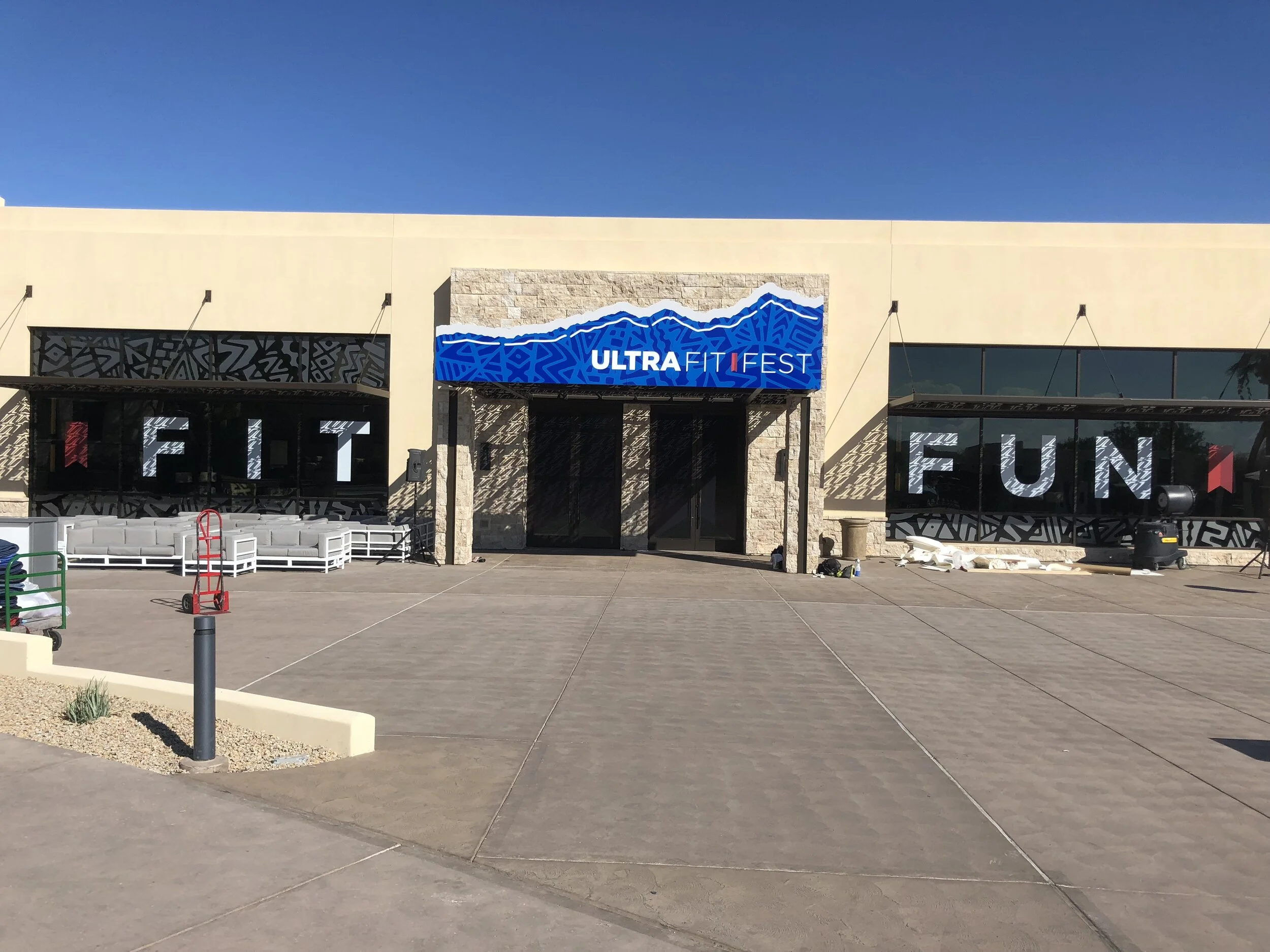 Front exterior of a building with a sign reading 'ULTRA FIT FEST' above the entrance, under a clear blue sky, with some furniture and equipment outside.